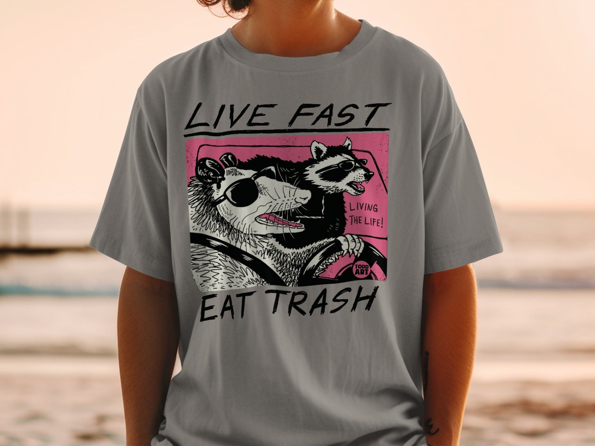 T-shirt with cartoon opossum and raccoon driving, "LIVE FAST EAT TRASH" text.