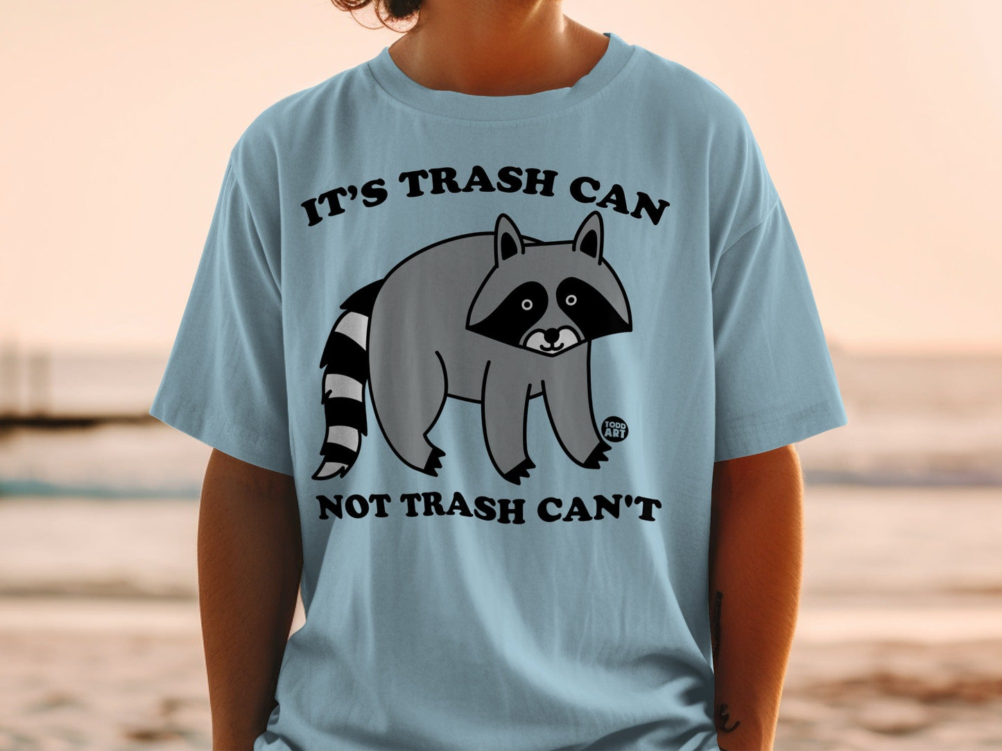Light blue t-shirt with raccoon graphic and text "IT'S TRASH CAN NOT TRASH CAN'T".