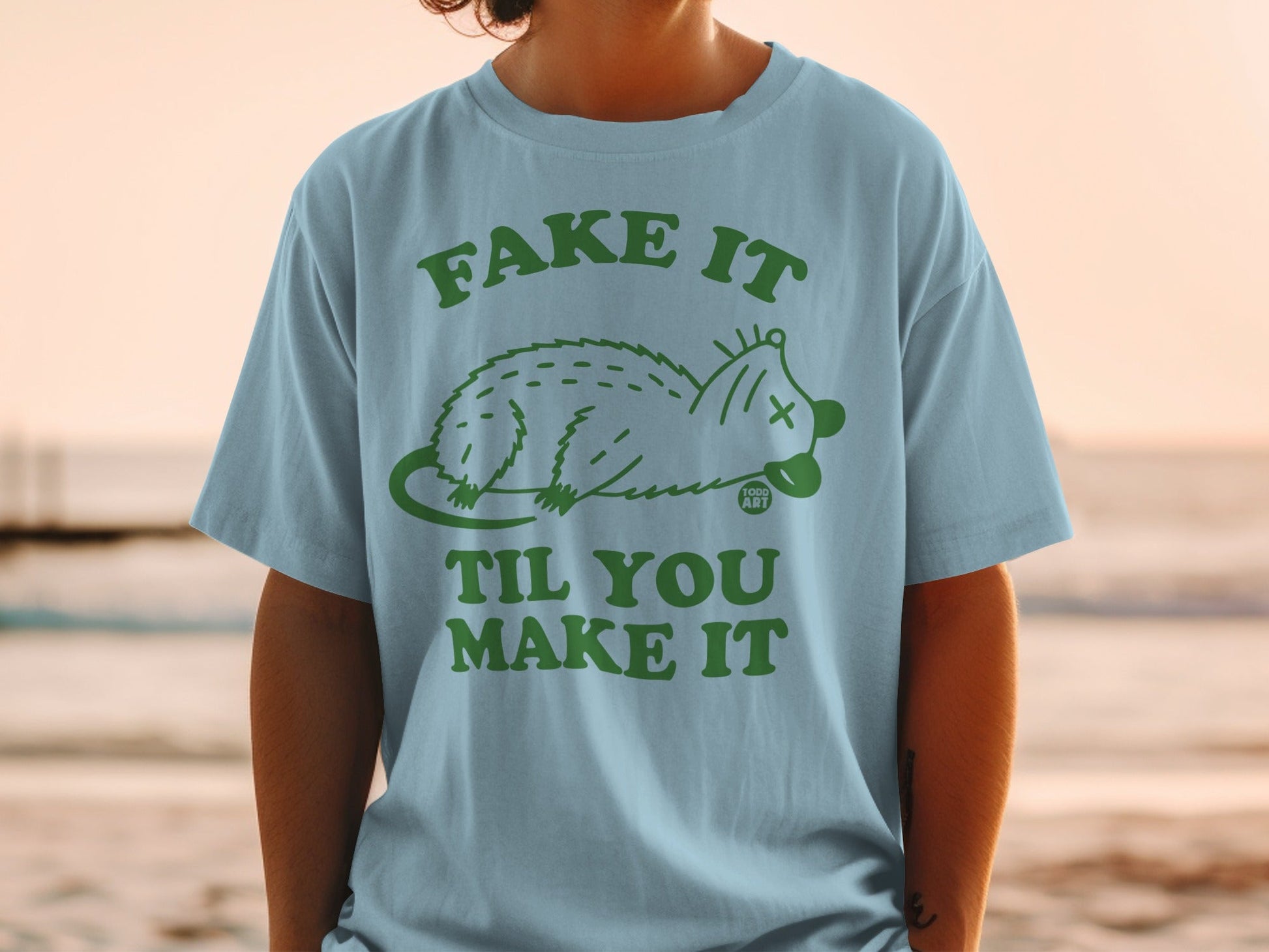 Light blue t-shirt with green text "FAKE IT TIL YOU MAKE IT" and a possum graphic.