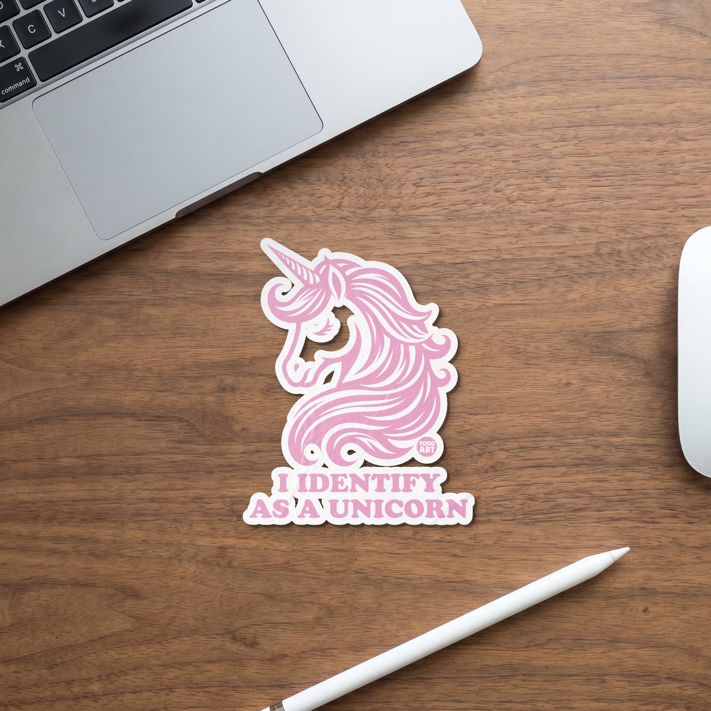 Pink unicorn sticker with text "I IDENTIFY AS A UNICORN" on wooden desk.