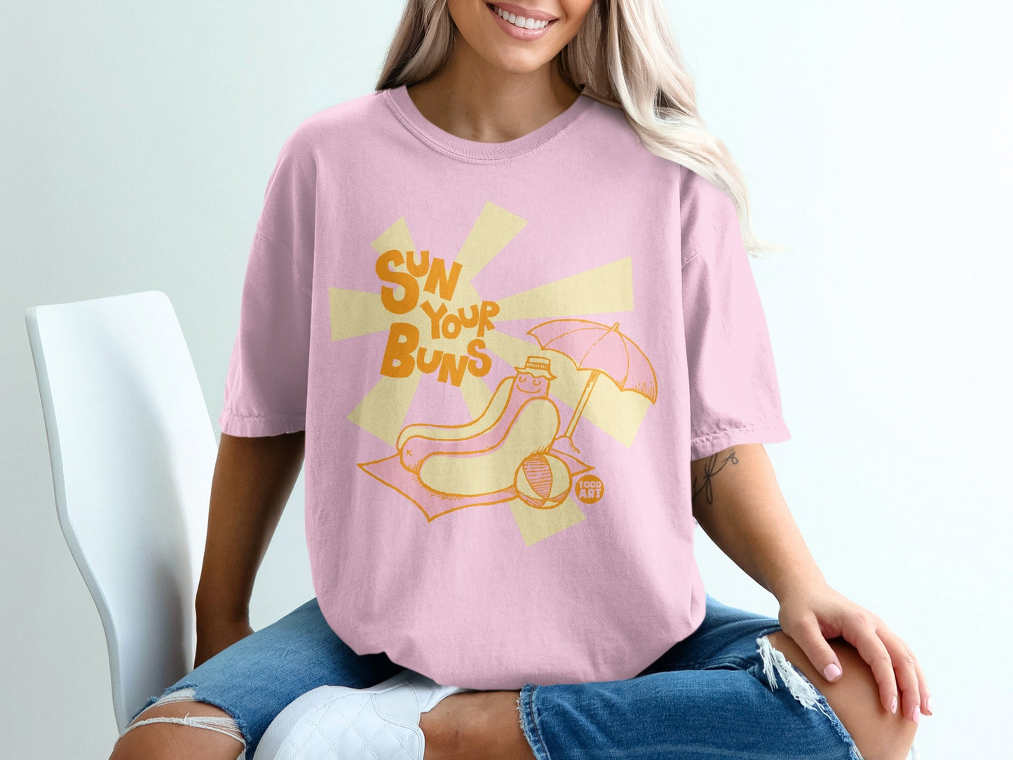 Pink t-shirt with "Sun Your Buns" text and cartoon hot dog on beach.