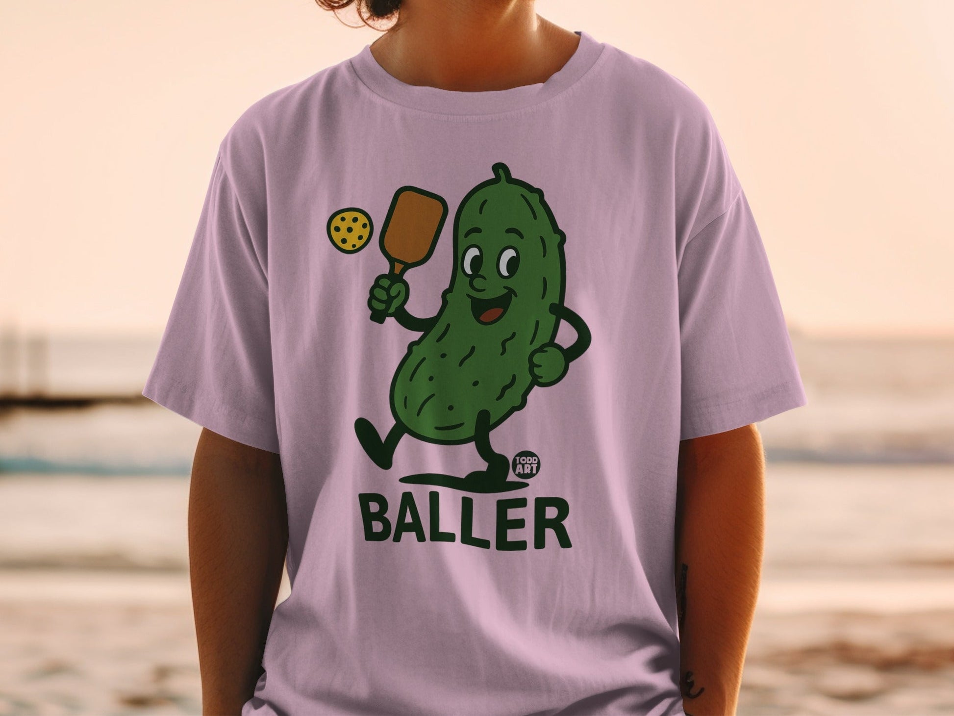 Cartoon pickle playing pickleball on a light purple t-shirt.