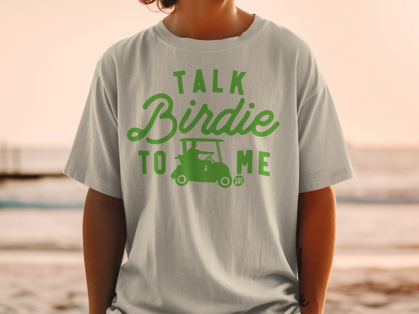Green text "Talk Birdie To Me" with golf cart graphic on a light gray t-shirt.