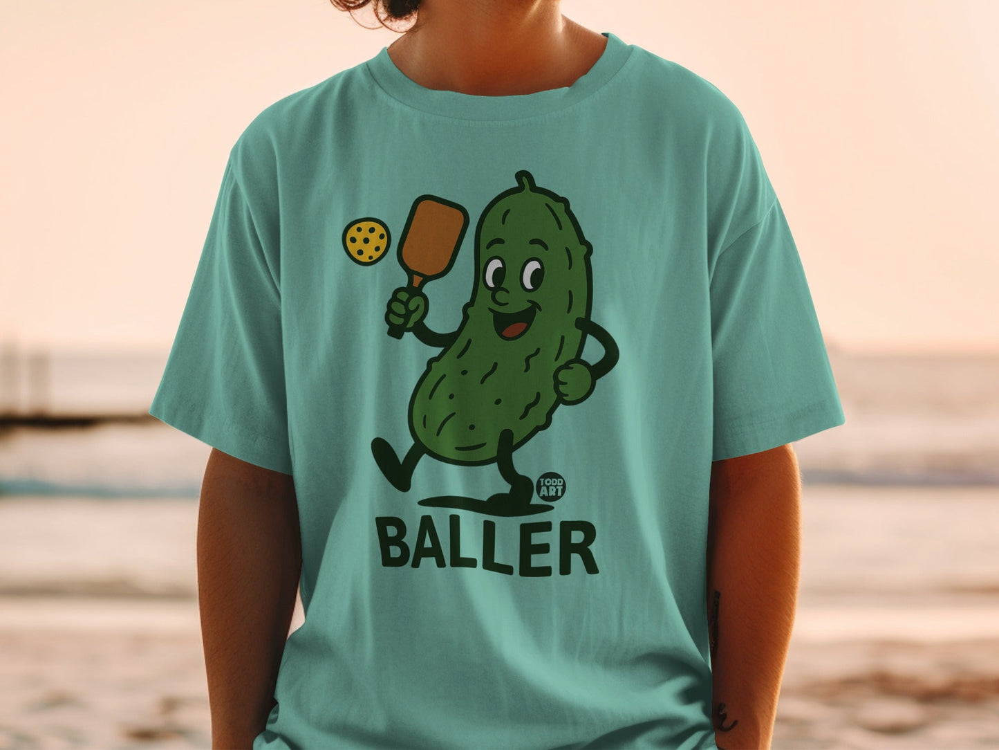 Green t-shirt with cartoon pickle playing pickleball, "Good Art" logo below.