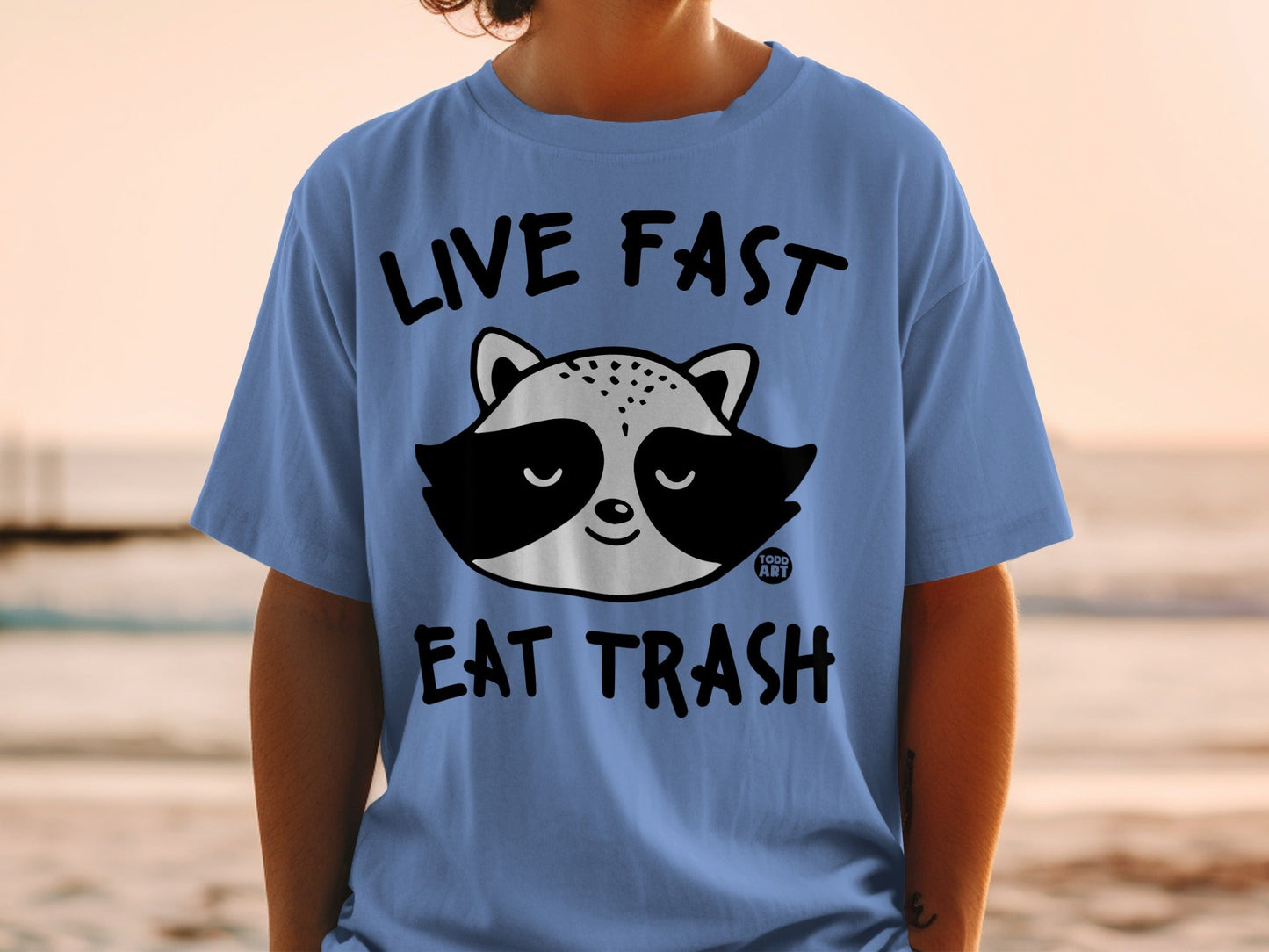 Light blue t-shirt with raccoon graphic and text: "LIVE FAST EAT TRASH".