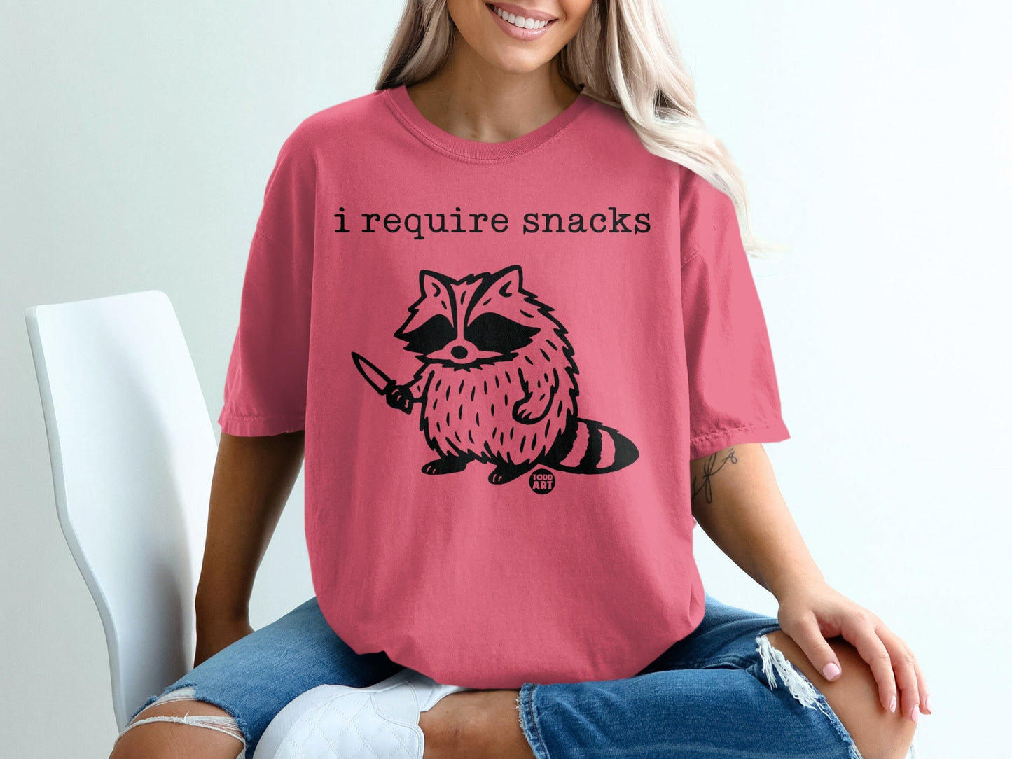 Pink t-shirt with raccoon holding a knife and the text "I require snacks".