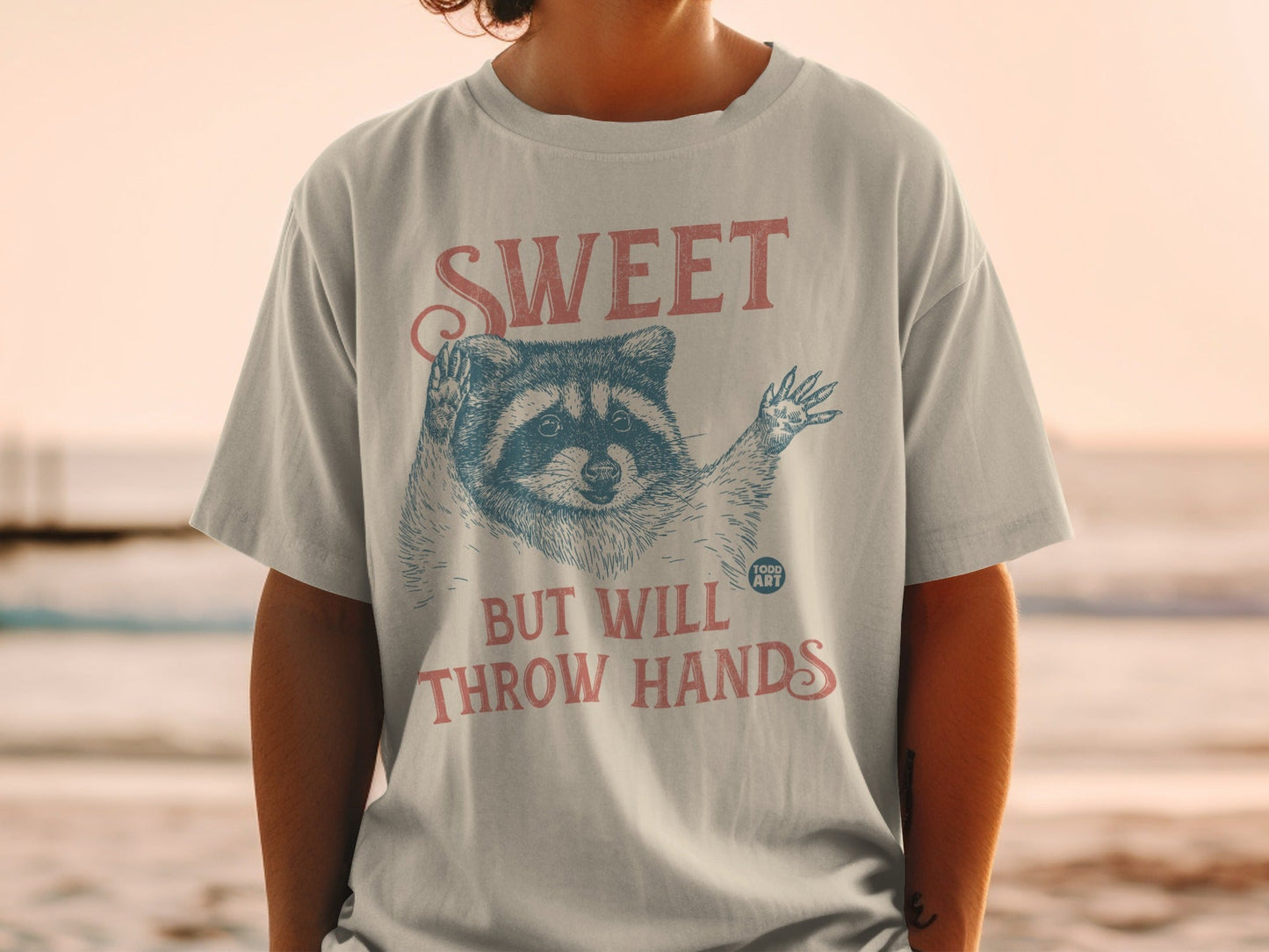 T-shirt with raccoon graphic and text "SWEET BUT WILL THROW HANDS".