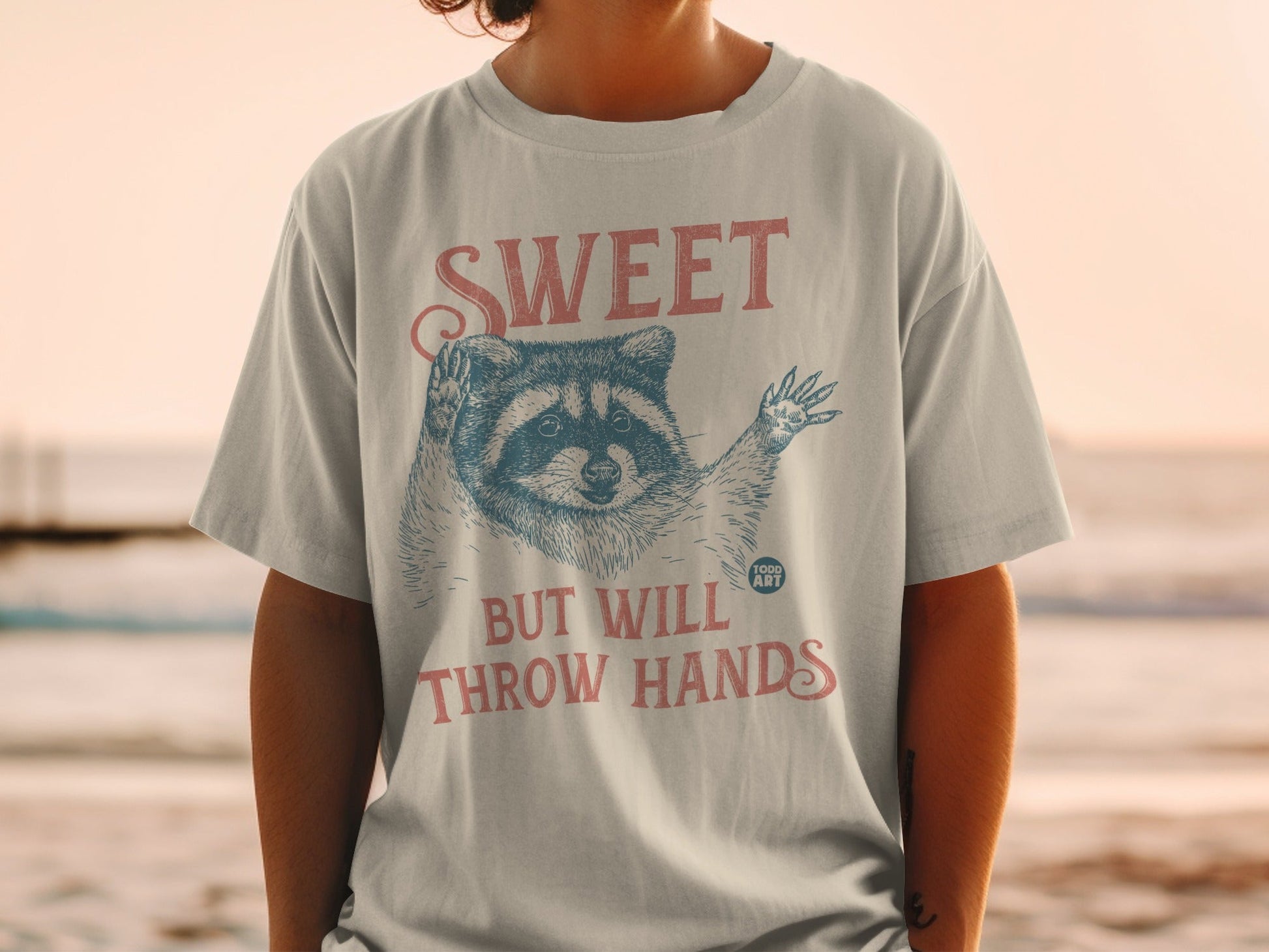 T-shirt with raccoon graphic and text "SWEET BUT WILL THROW HANDS".