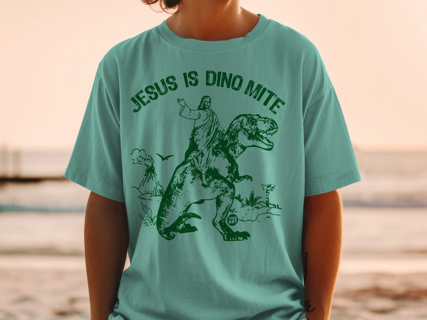 Teal t-shirt with graphic of Jesus riding a T-Rex, "Jesus is Dino Mite" text.