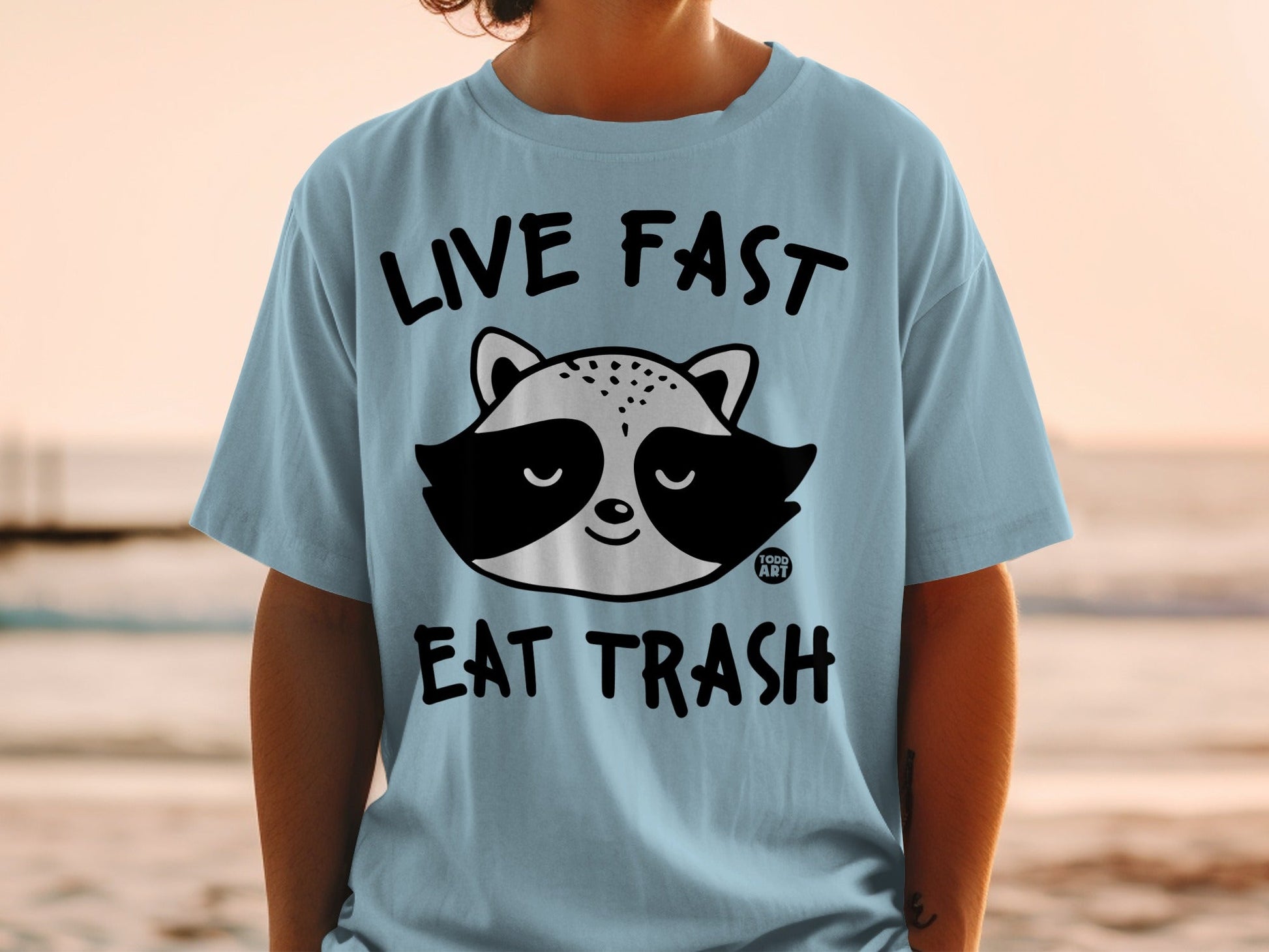 Light blue t-shirt with a raccoon graphic and "Live Fast Eat Trash" text.