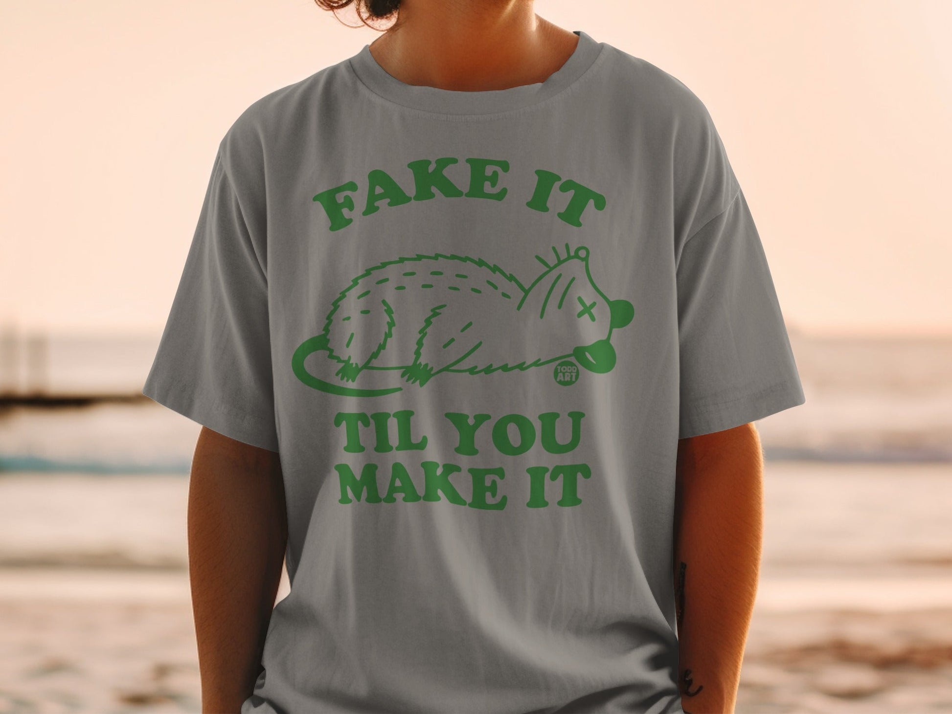 Grey t-shirt with green text "Fake It Til You Make It" and a cartoon opossum.
