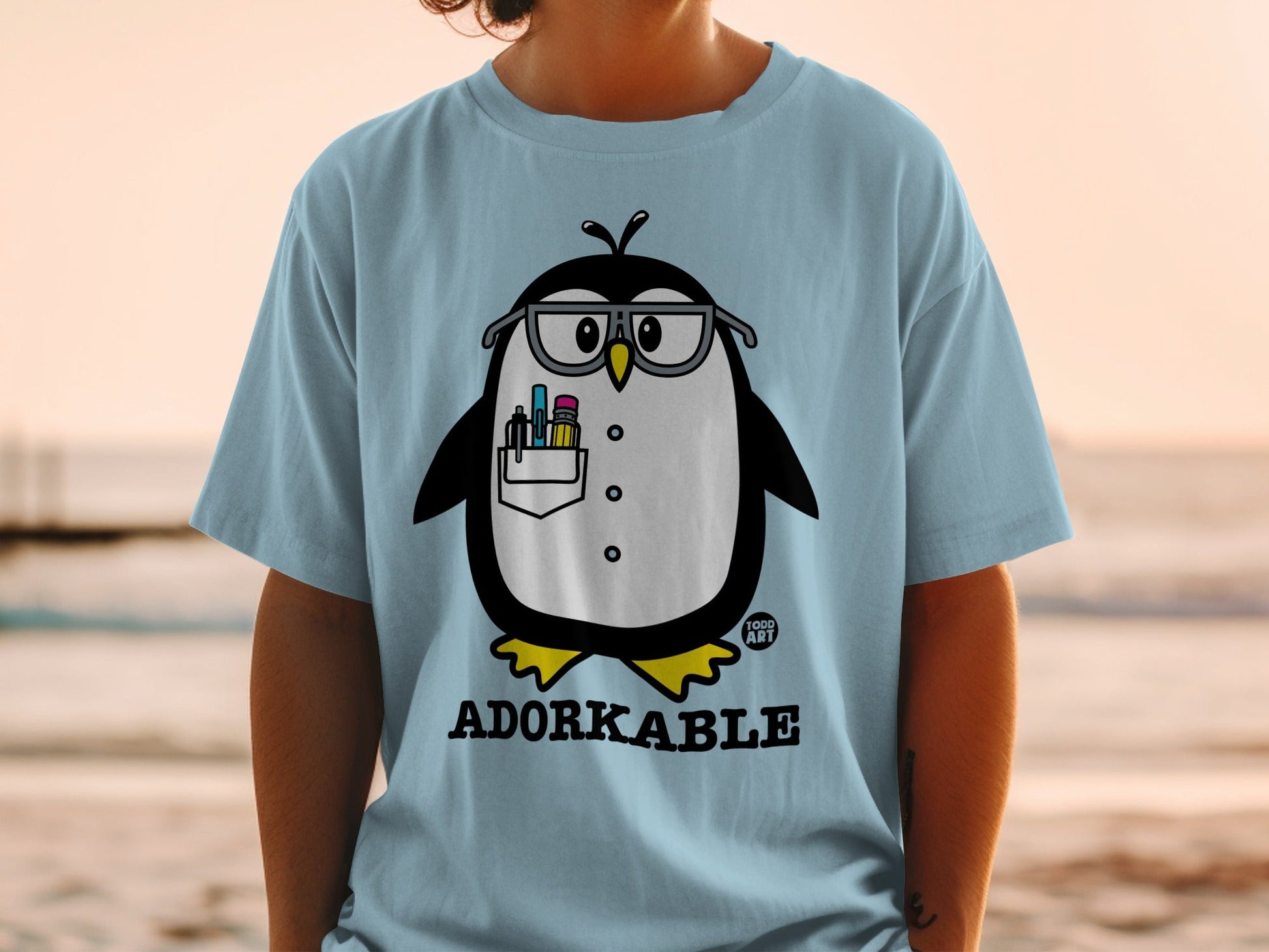 Light blue t-shirt with a cartoon penguin wearing glasses and holding art supplies.