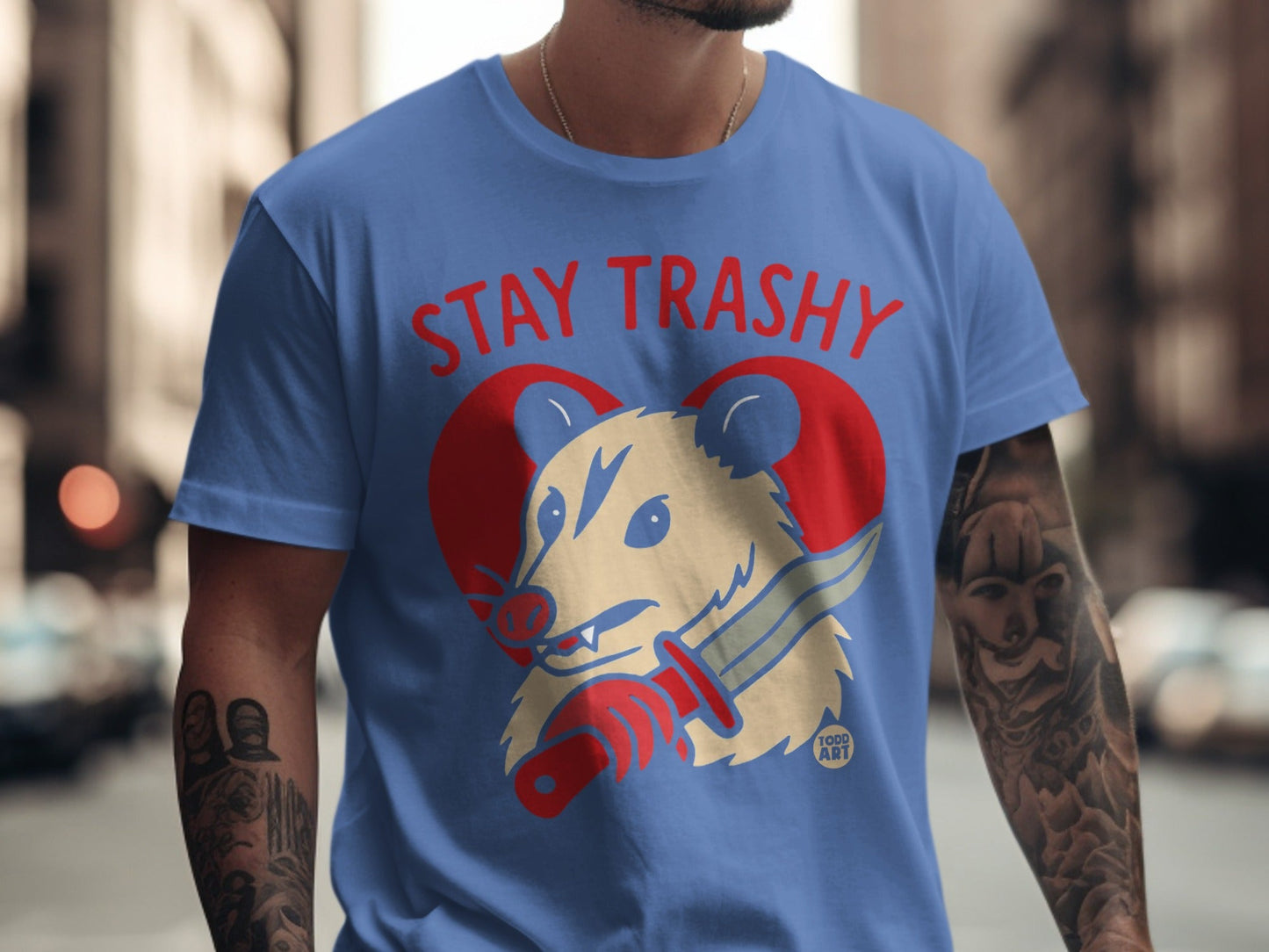 Blue t-shirt with "STAY TRASHY" text and cartoon opossum holding a knife.