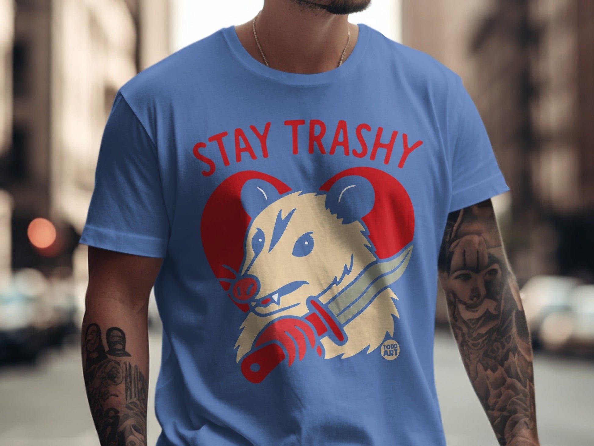 Blue t-shirt with "STAY TRASHY" text and cartoon opossum holding a knife.