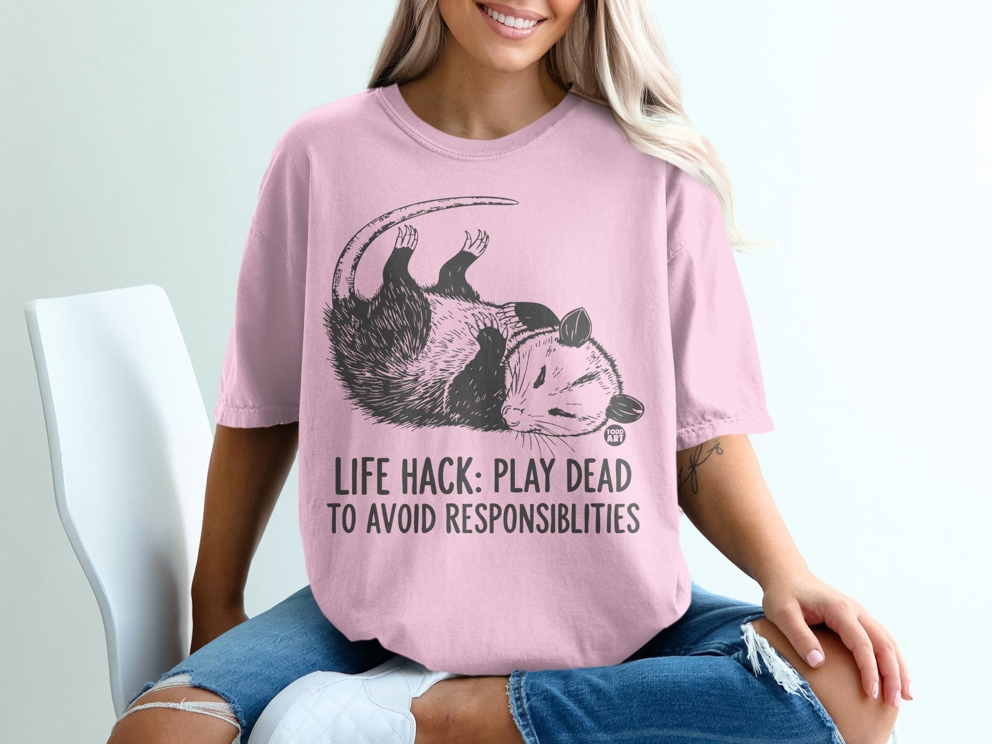 Pink t-shirt with opossum graphic and text "LIFE HACK: PLAY DEAD TO AVOID RESPONSIBILITIES".