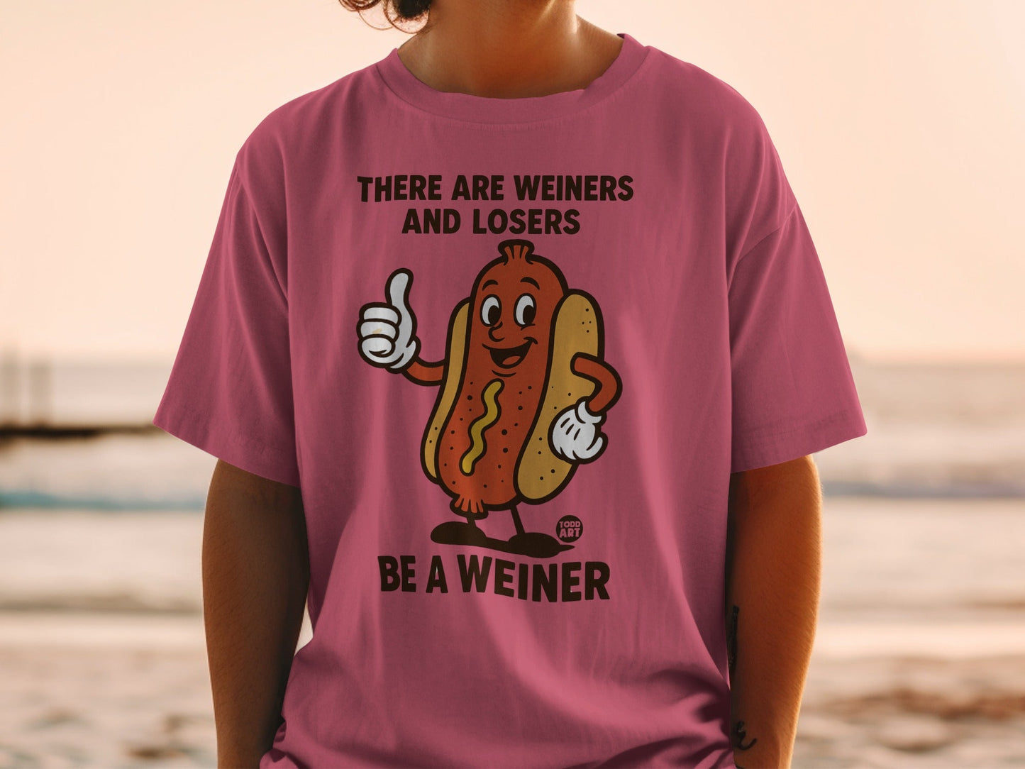 Pink t-shirt with a cartoon hot dog giving a thumbs up saying "BE A WEINER".
