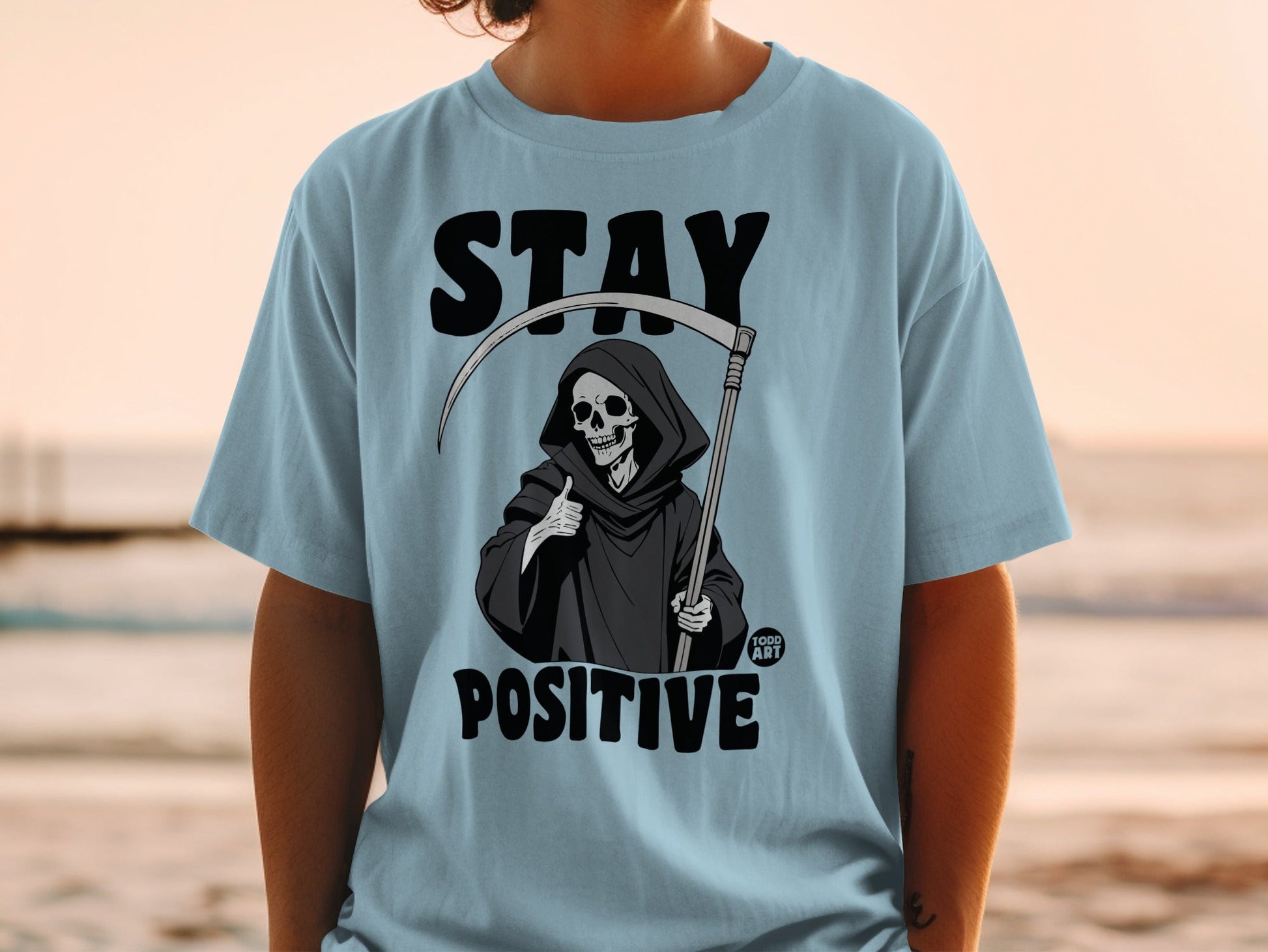 Grim reaper giving thumbs up on light blue t-shirt with "STAY POSITIVE" text.