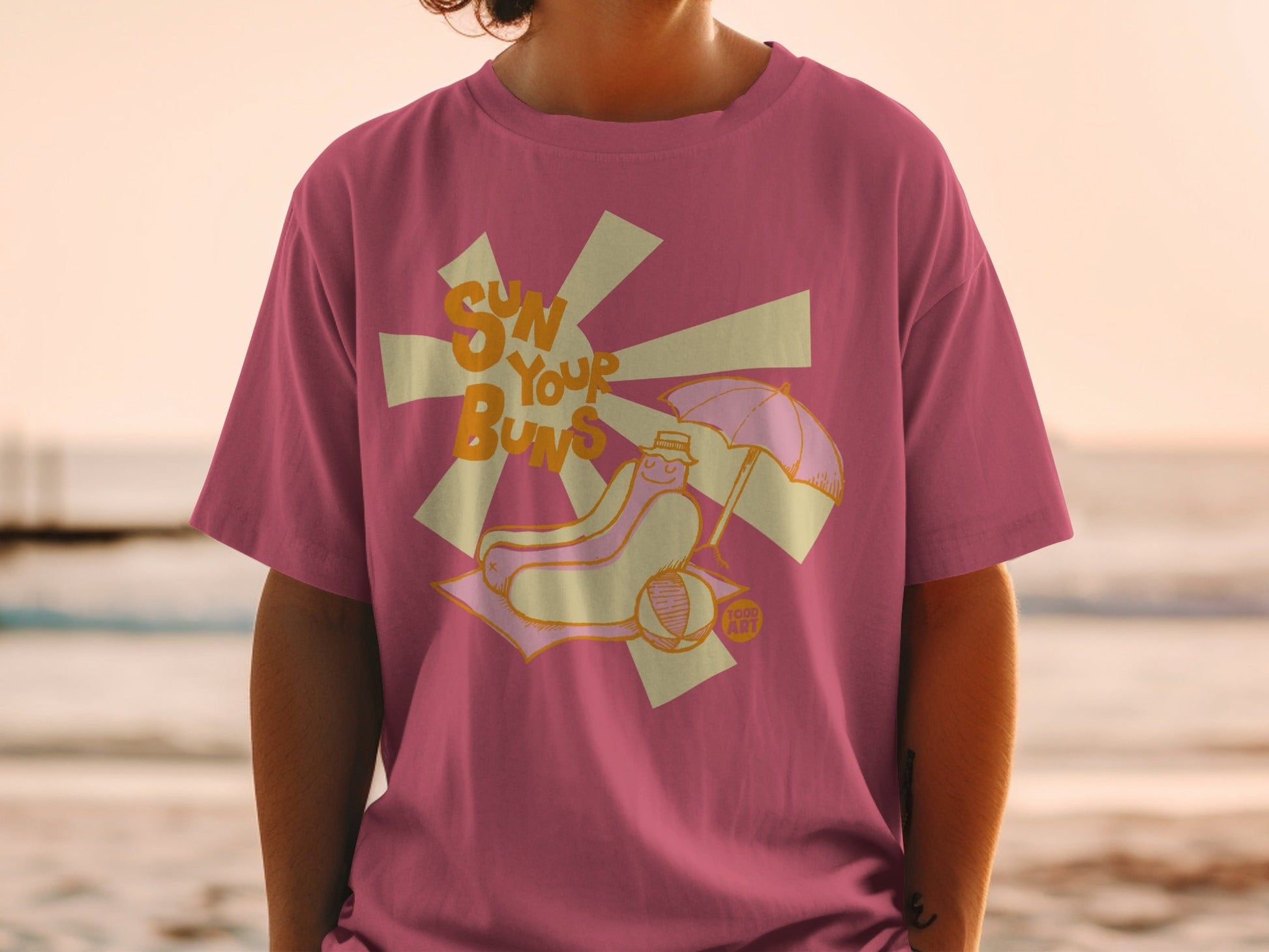 Pink t-shirt with cartoon hot dog wearing sunglasses and hat at beach.