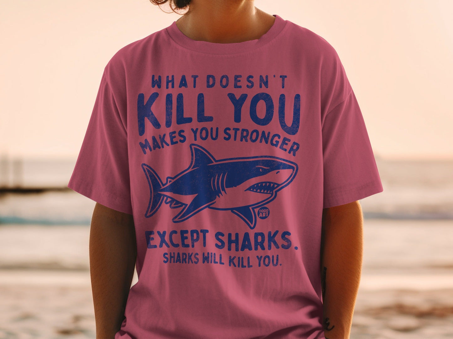 Pink t-shirt with shark graphic and "What doesn't kill you makes you stronger, except sharks" text.