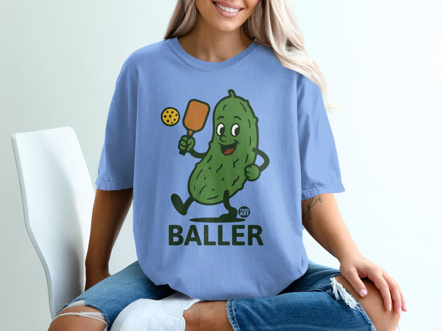 Blue t-shirt with cartoon pickle playing pickleball, text reads BALLER GOOD ART.