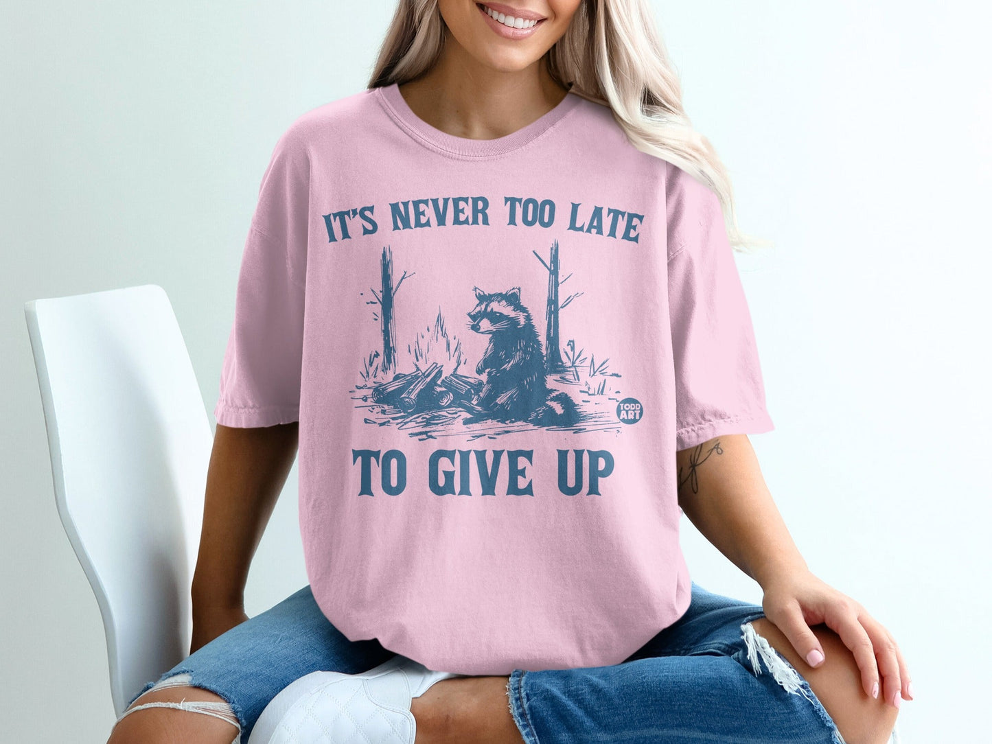 Pink t-shirt with raccoon and campfire graphic, text "IT'S NEVER TOO LATE TO GIVE UP".