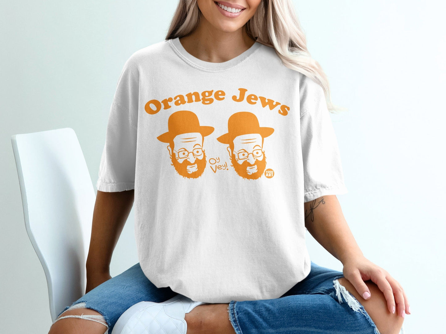 White t-shirt with "Orange Jews" and cartoon faces, sitting on a chair.