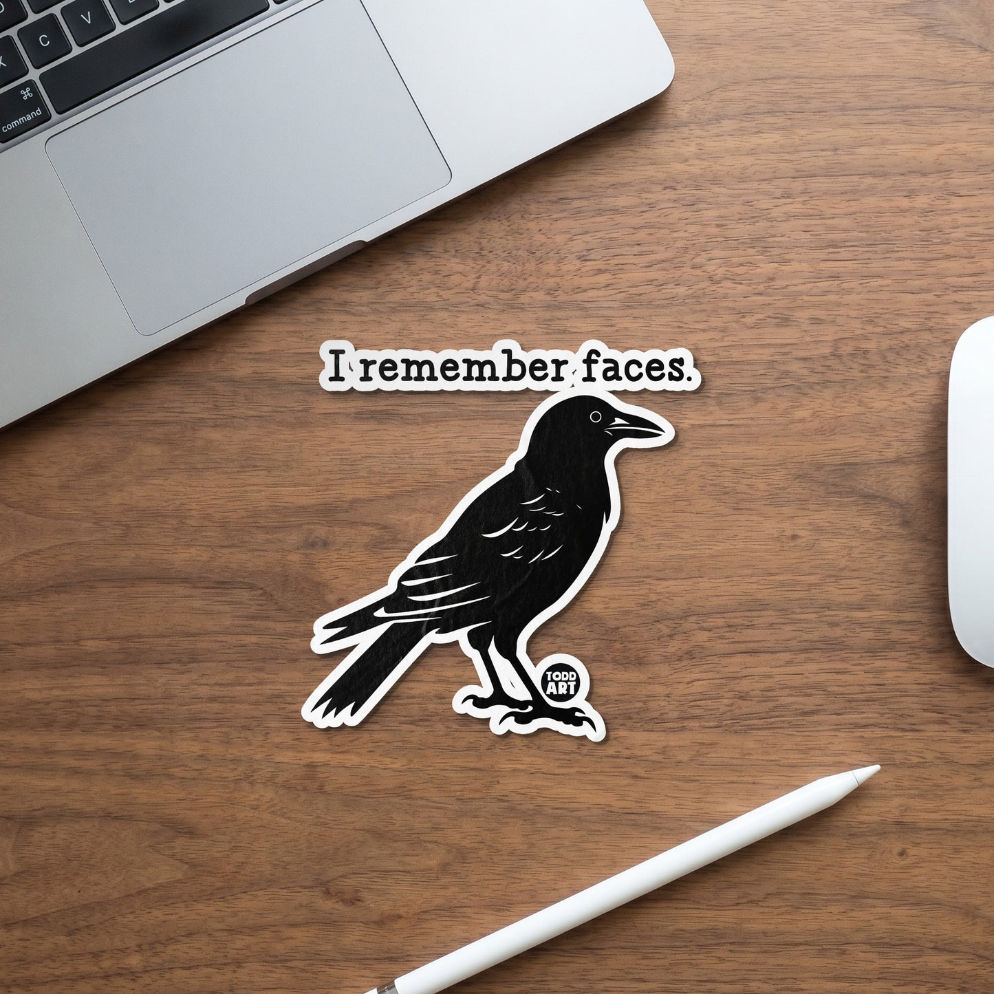 Black crow sticker with "I remember faces." text on a wooden desk with laptop and mouse.