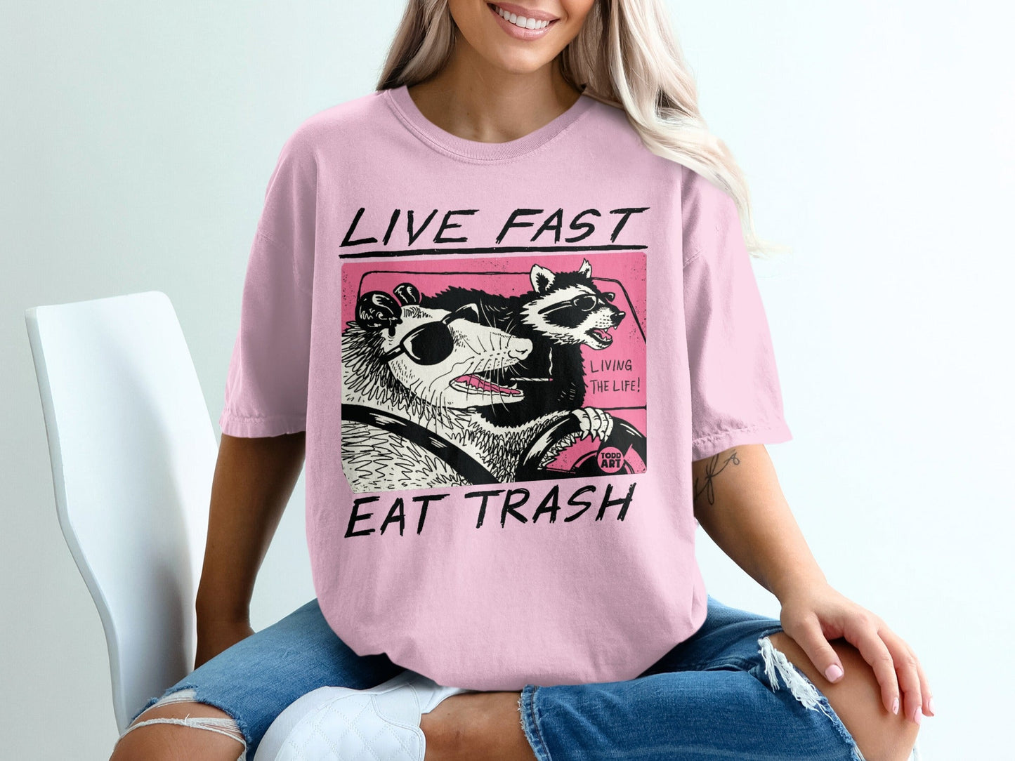 Pink t-shirt with raccoon and possum wearing sunglasses driving a car.