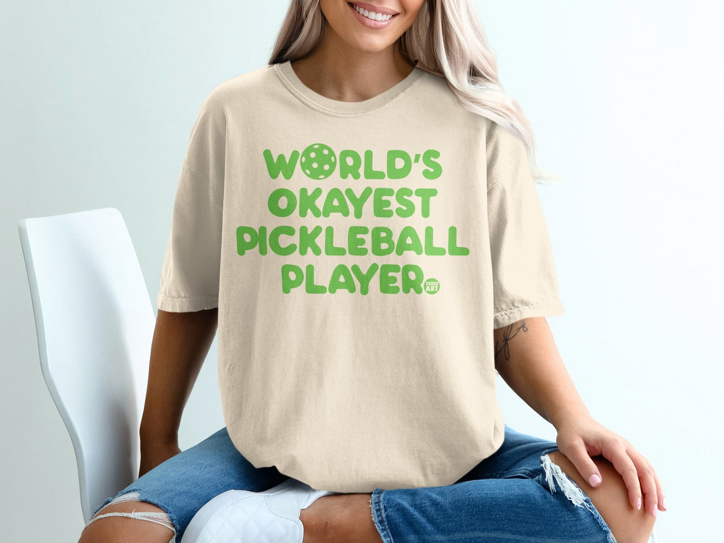 Cream t-shirt with "World's Okayest Pickleball Player" text and pickleball graphic.