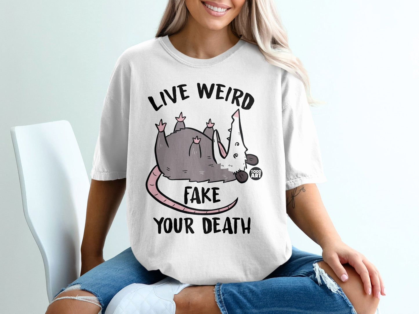 White t-shirt with "LIVE WEIRD FAKE YOUR DEATH" text and a cartoon possum.