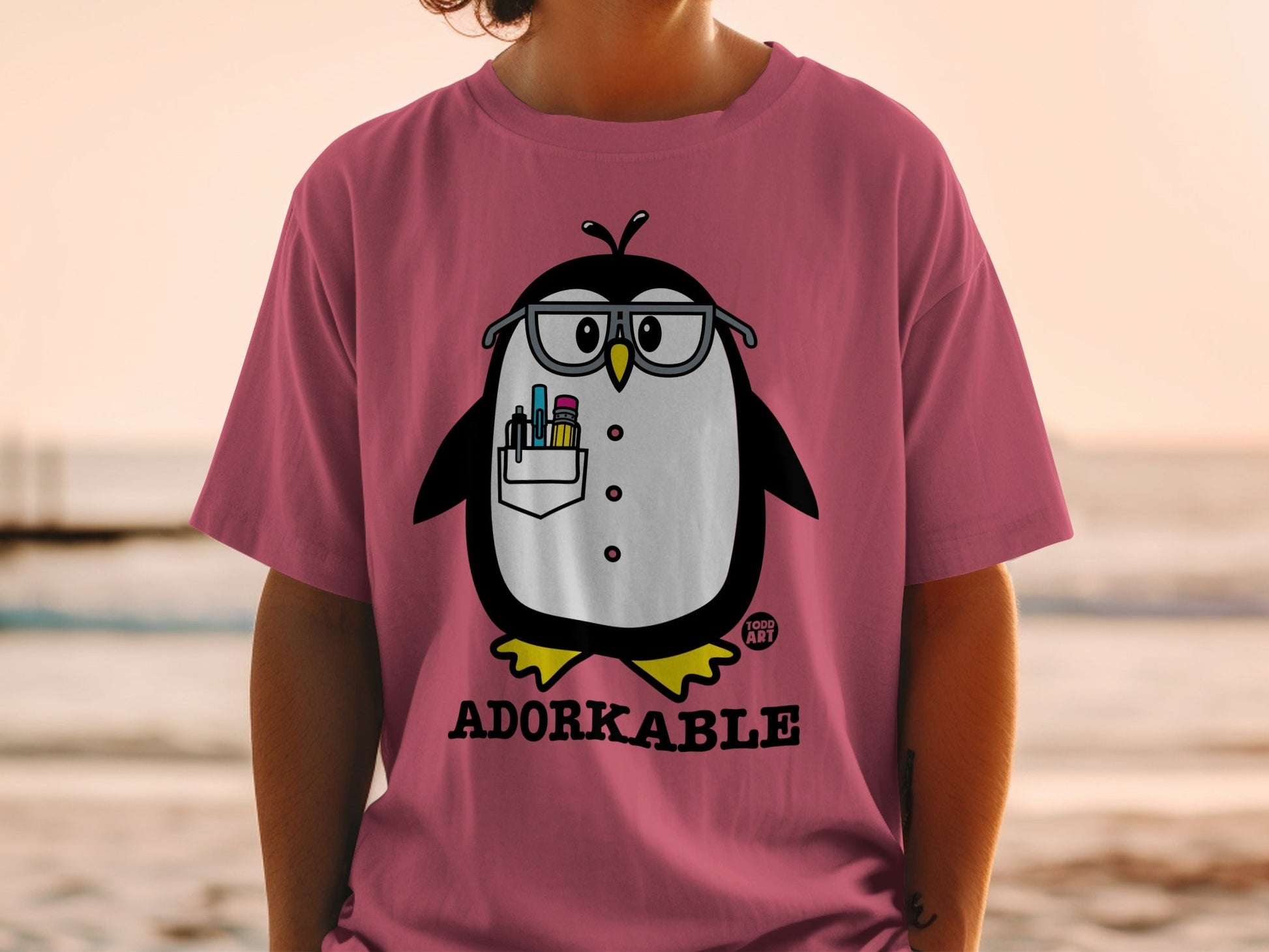 Pink t-shirt with a cartoon penguin wearing glasses and holding pens.