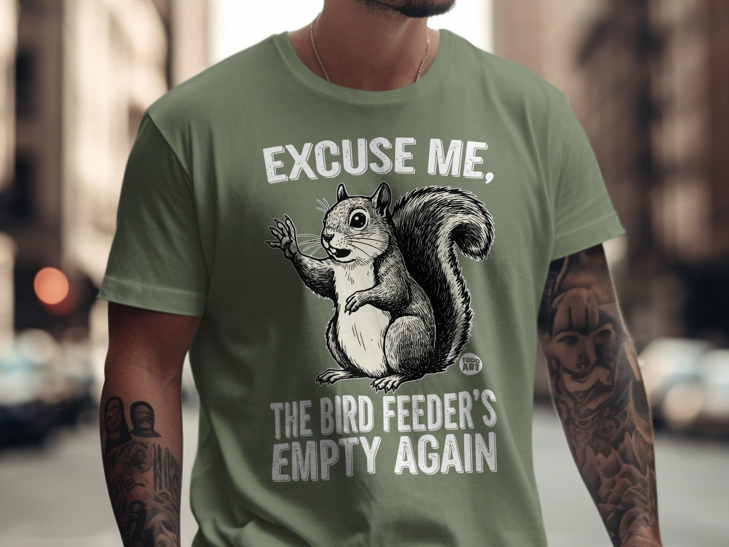 Squirrel on olive green t-shirt says "Excuse me, the bird feeder's empty again."