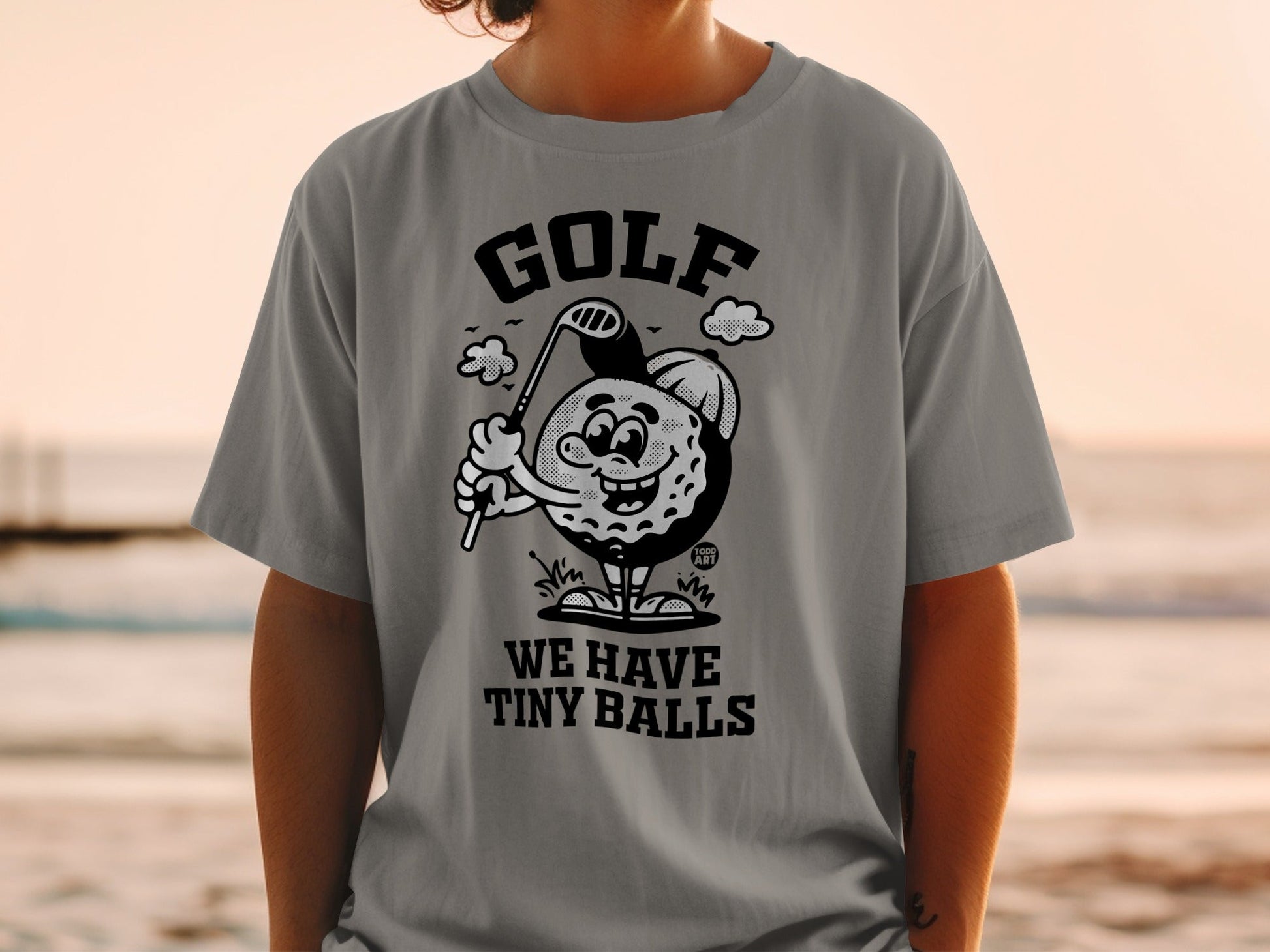 Gray t-shirt with cartoon golf ball holding club, text "WE HAVE TINY BALLS".