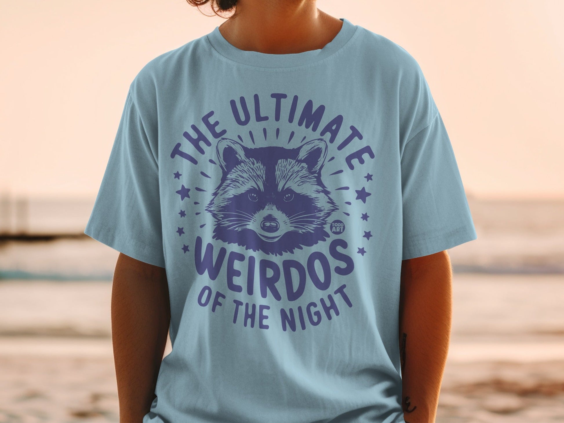 Light blue t-shirt with a purple raccoon illustration and text "THE ULTIMATE WEIRDOS OF THE NIGHT".