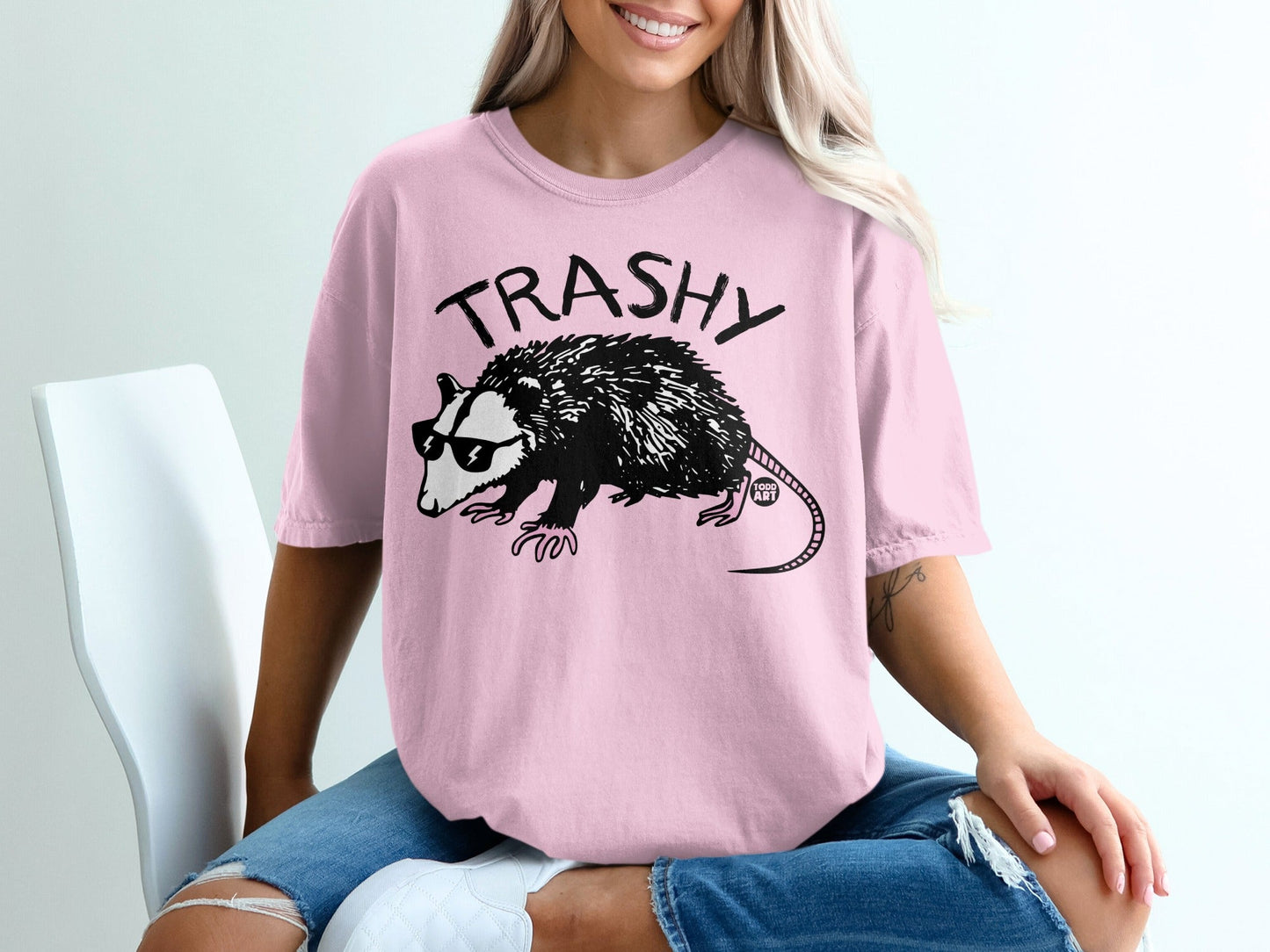 Pink t-shirt with "TRASHY" text and a cartoon opossum wearing sunglasses.