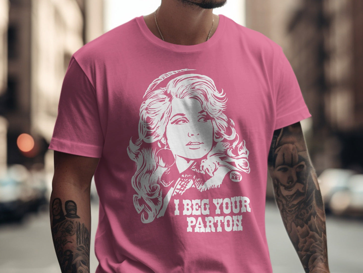 Pink t-shirt featuring a white graphic of Dolly Parton and "I BEG YOUR PARTON".
