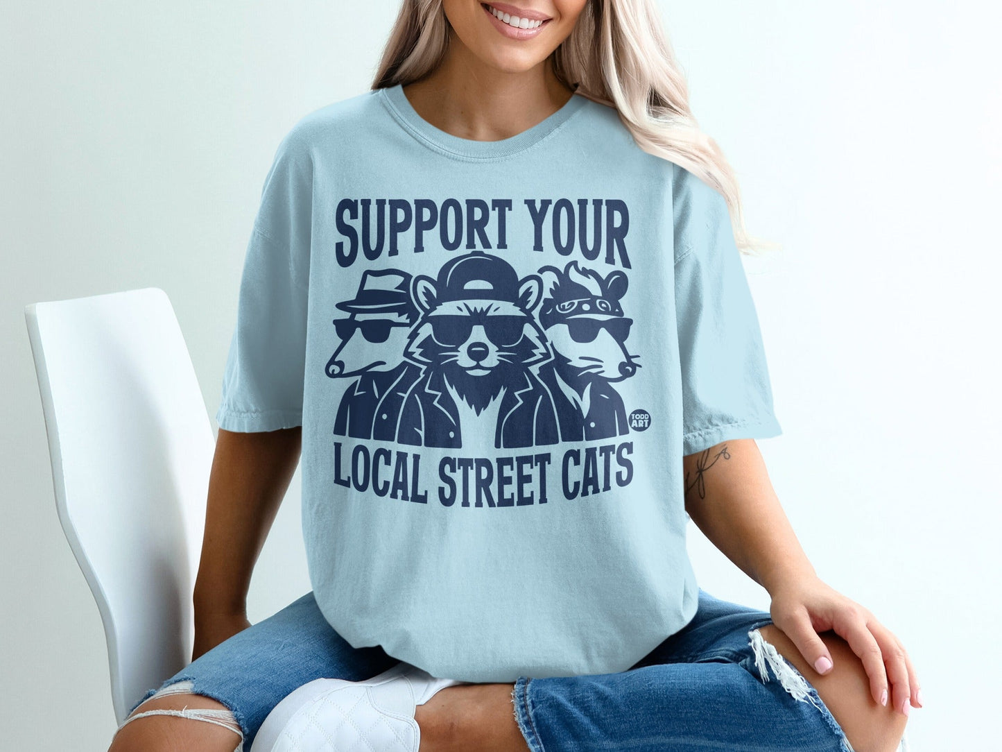 Light blue t-shirt with dark blue graphic of cool cats wearing sunglasses and hats.