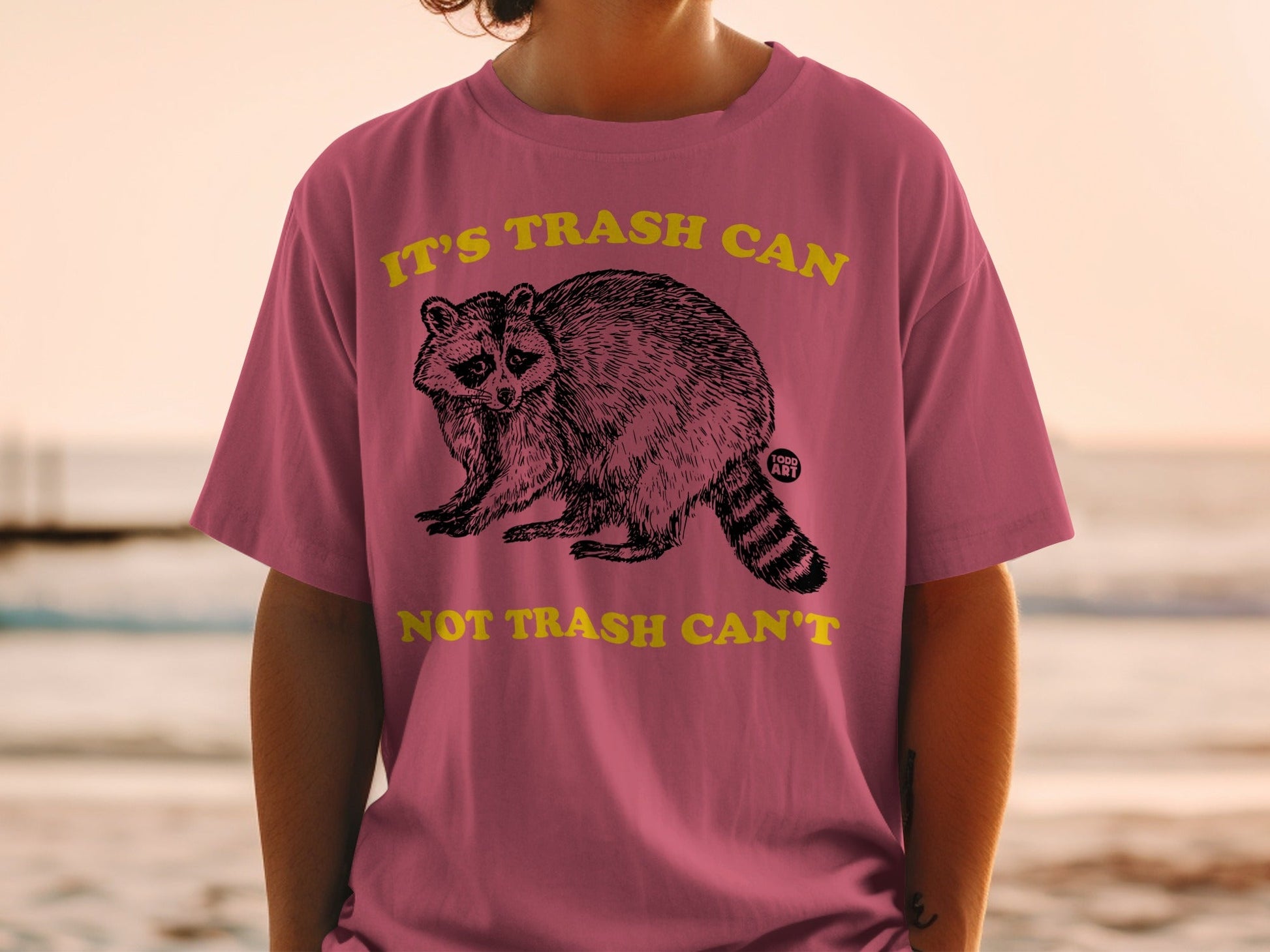 Pink t-shirt with raccoon graphic and text "IT'S TRASH CAN NOT TRASH CAN'T".