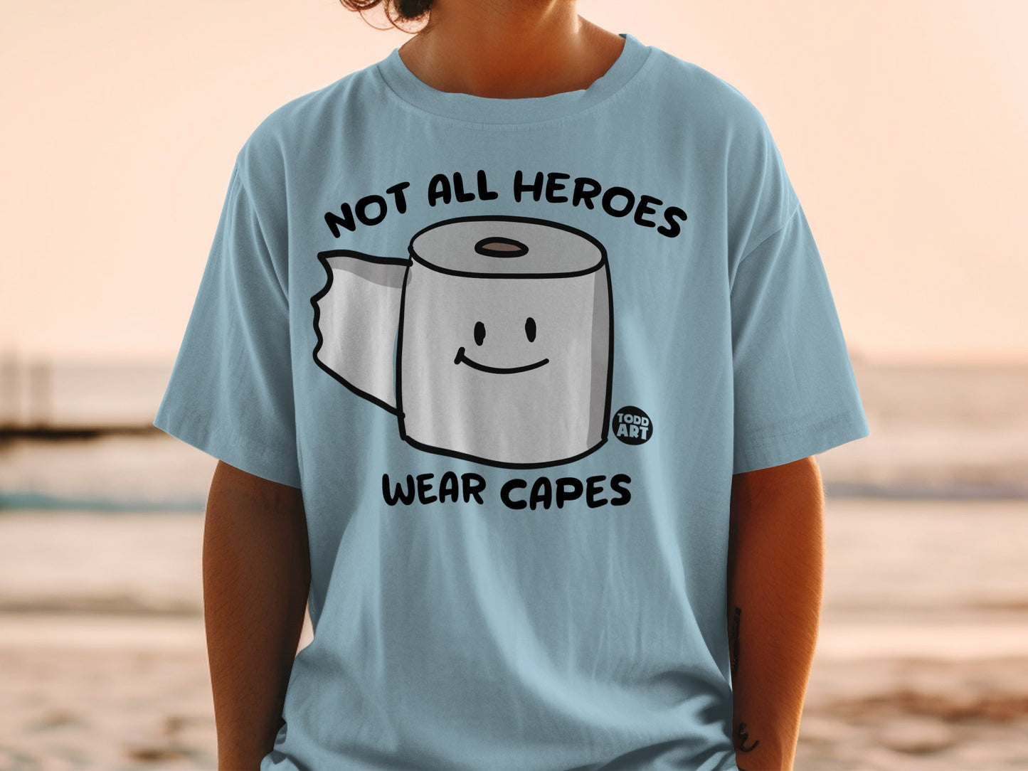 Light blue t-shirt with a smiling toilet paper roll and "Not All Heroes Wear Capes" text.