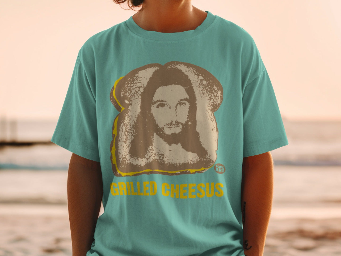 Teal t-shirt featuring grilled cheese sandwich with Jesus portrait and "GRILLED CHEESUS" text.