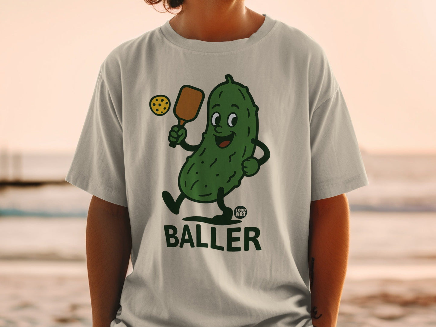T-shirt featuring a smiling pickle character holding a pickleball paddle and ball.