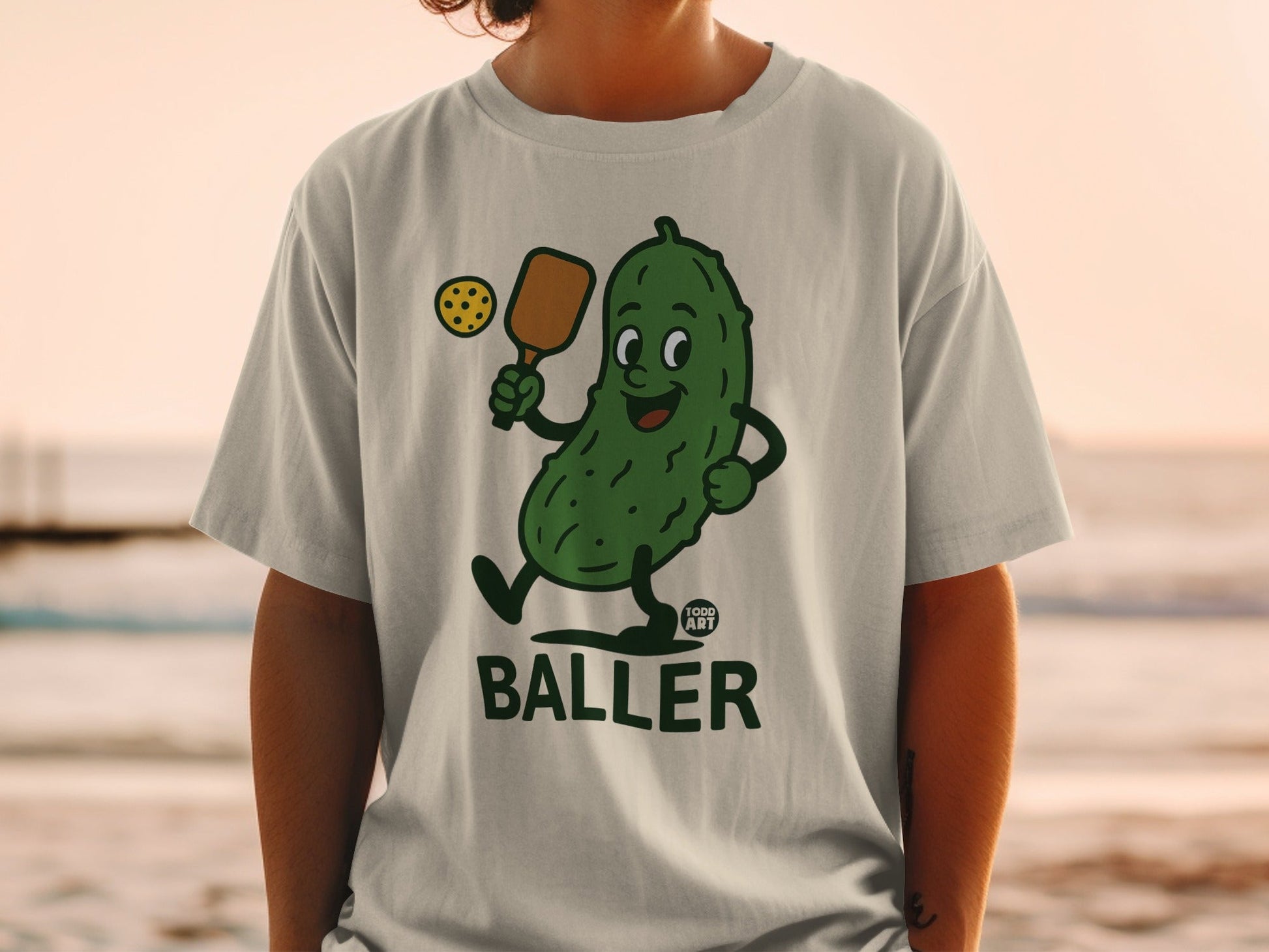 T-shirt featuring a smiling pickle character holding a pickleball paddle and ball.