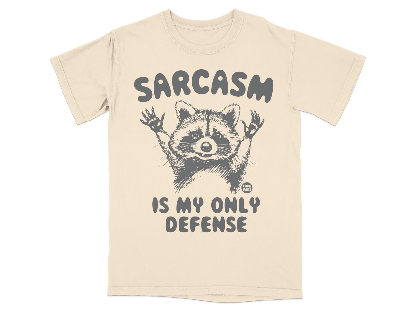 Cream t-shirt with raccoon graphic and "Sarcasm is my only defense" text.