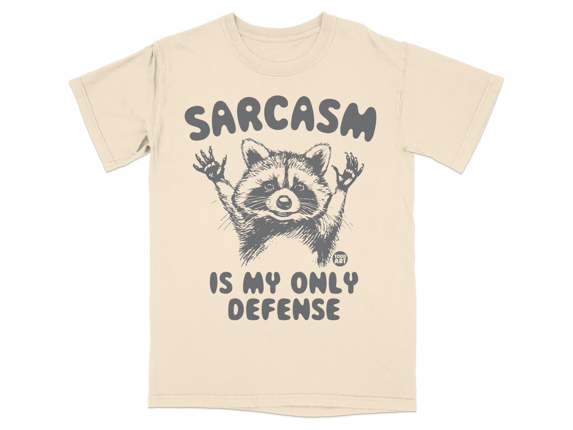 Cream t-shirt with raccoon graphic and "Sarcasm is my only defense" text.