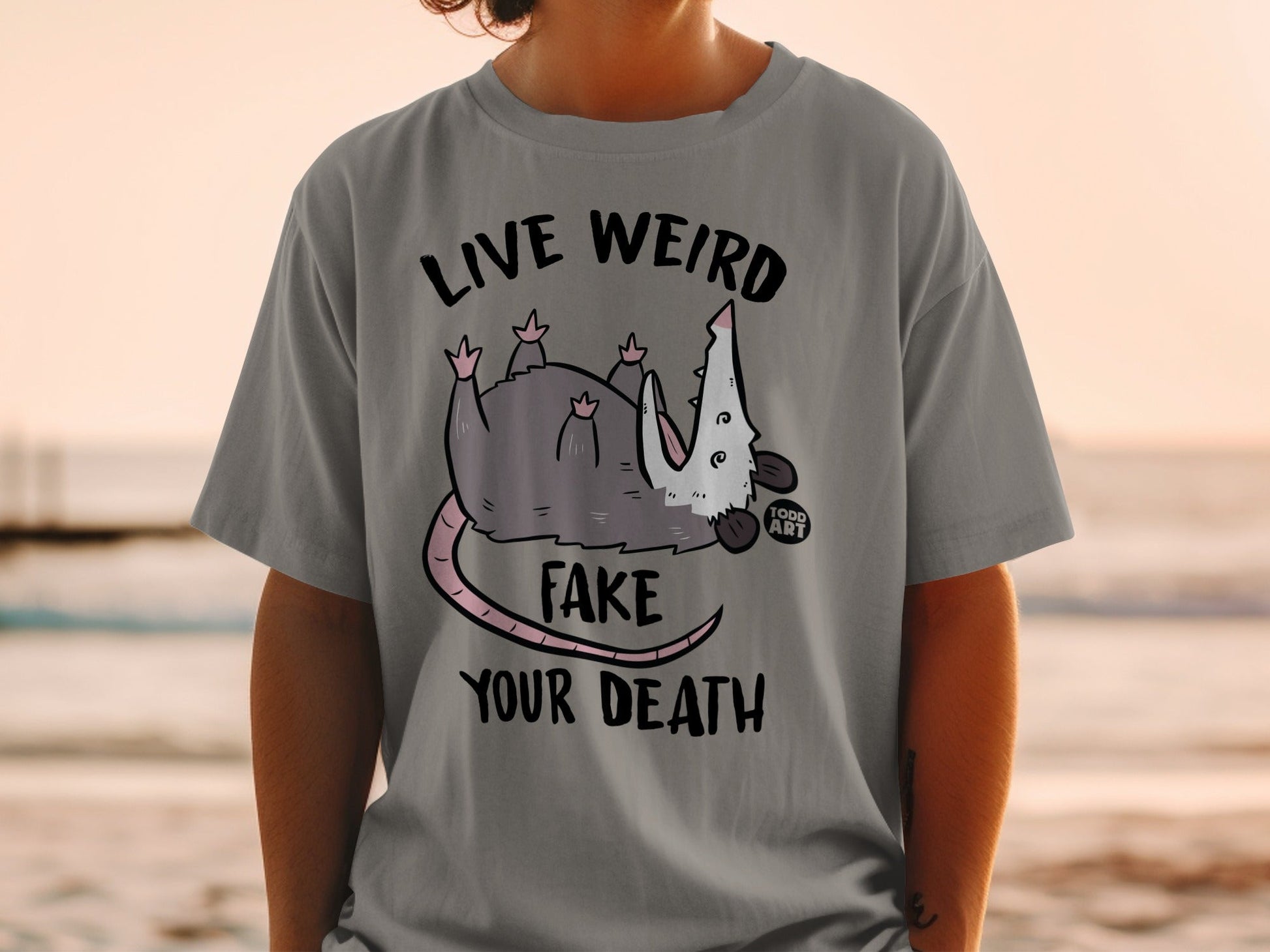 Grey t-shirt with a possum graphic and text: LIVE WEIRD FAKE YOUR DEATH.