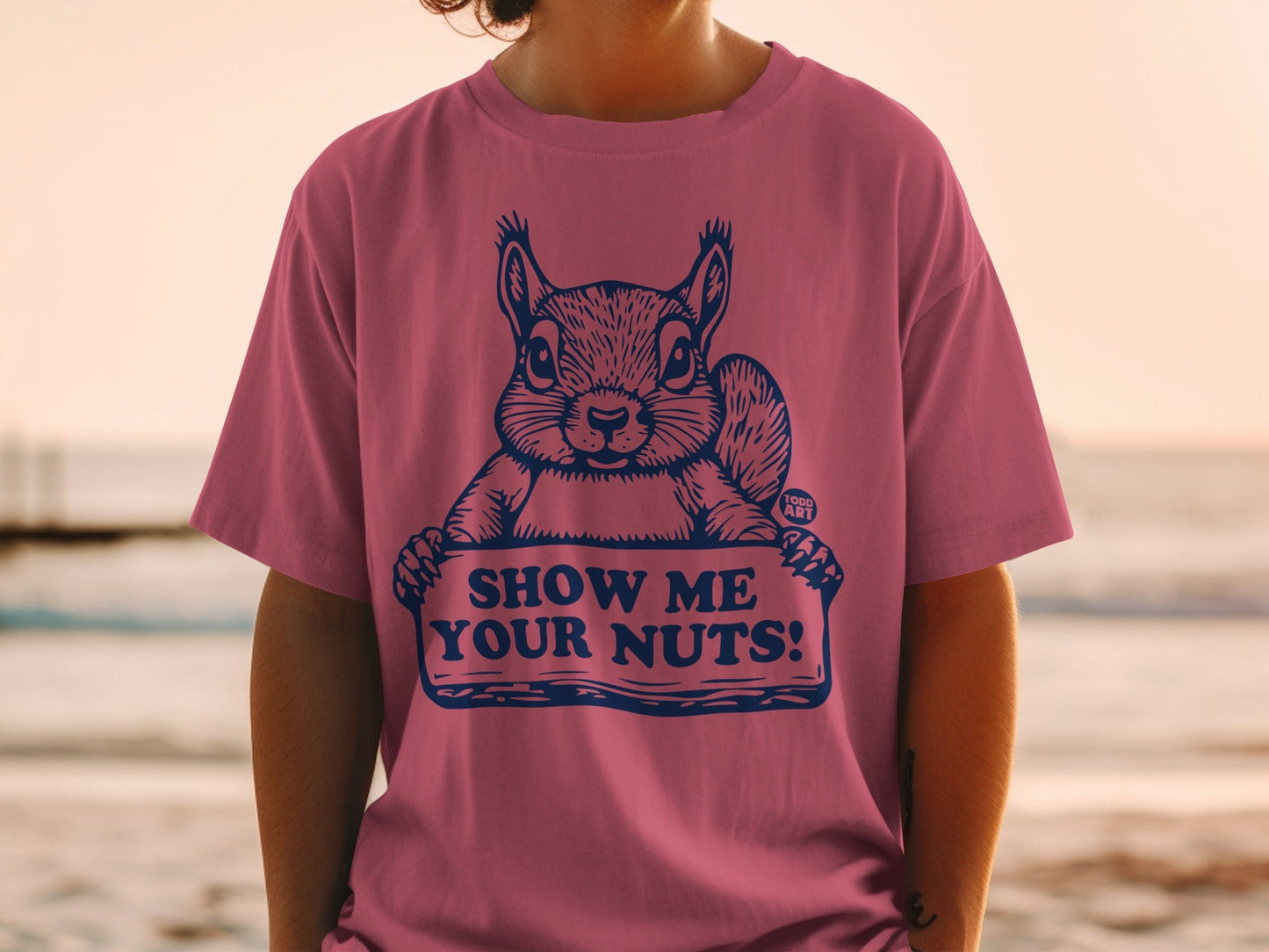 Pink t-shirt with a squirrel holding a sign that says "SHOW ME YOUR NUTS!".