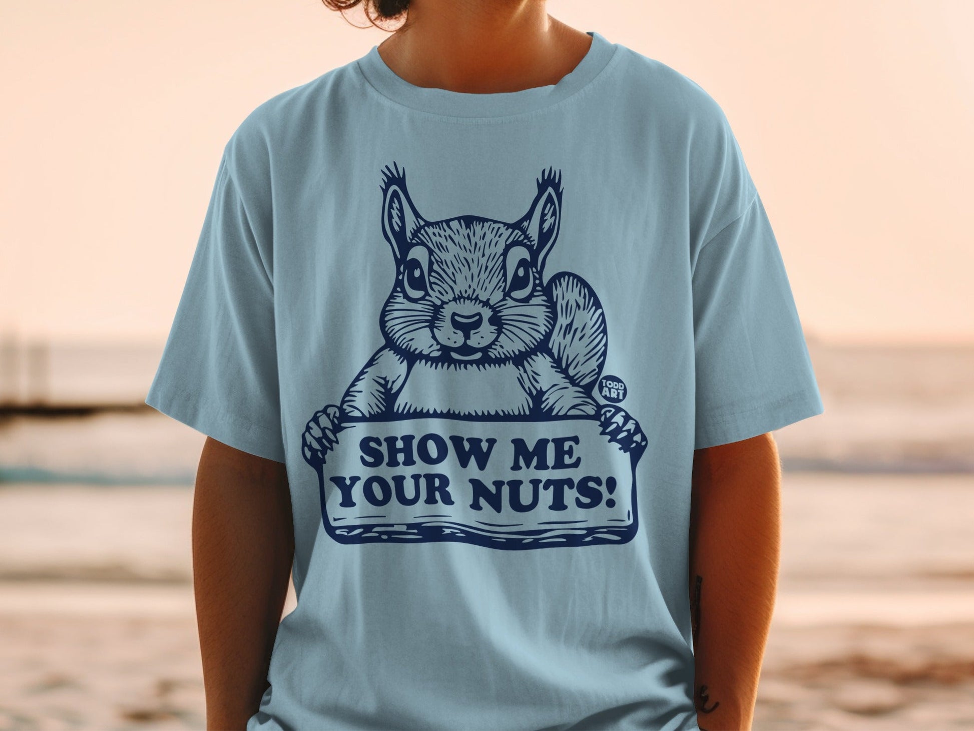 Light blue t-shirt with a cartoon squirrel holding a sign that reads "SHOW ME YOUR NUTS!".