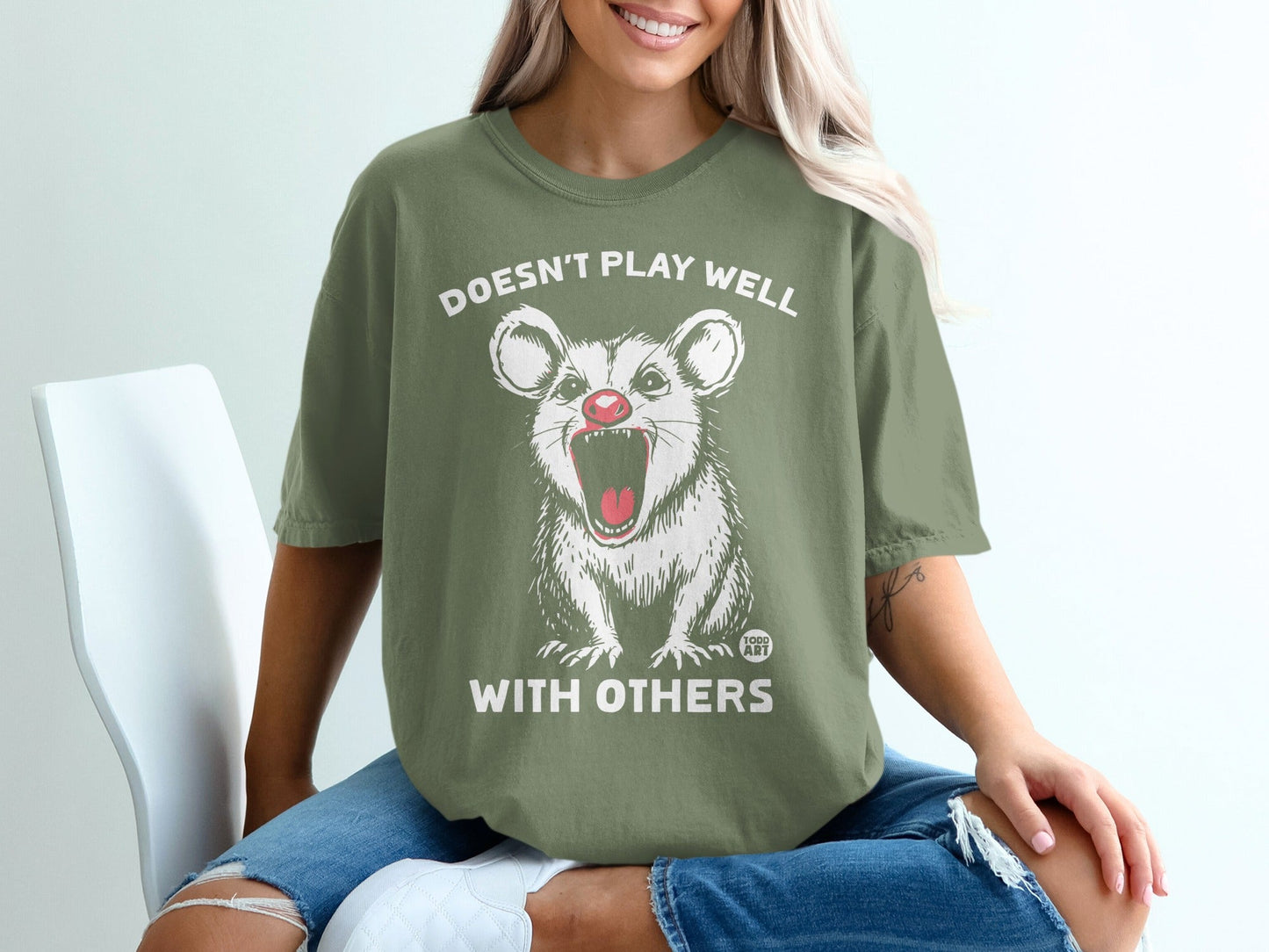 Green t-shirt with a cartoon opossum graphic and text "DOESN'T PLAY WELL WITH OTHERS".