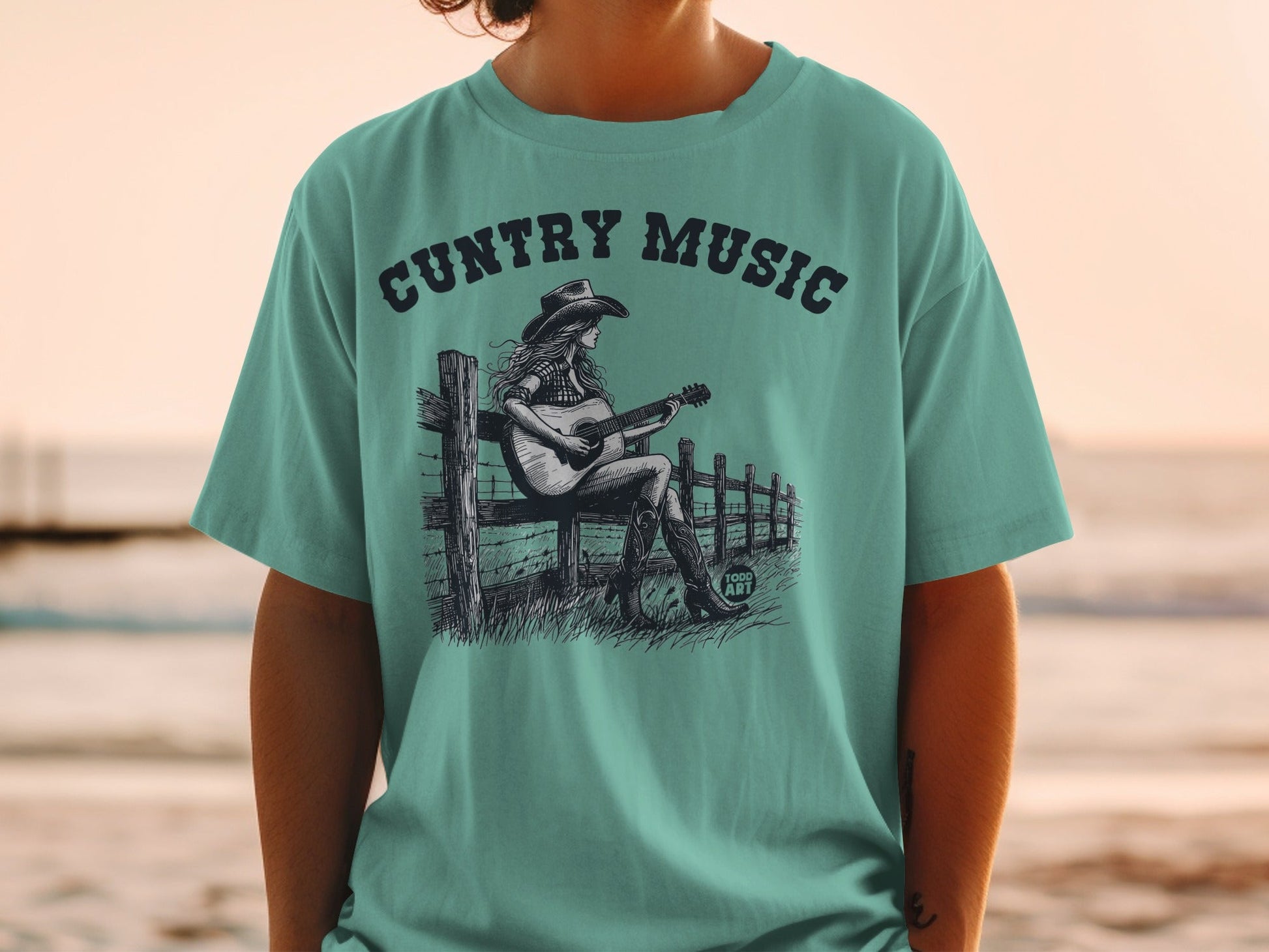 Teal t-shirt with "CUNTry Music" and cowboy playing guitar on fence.