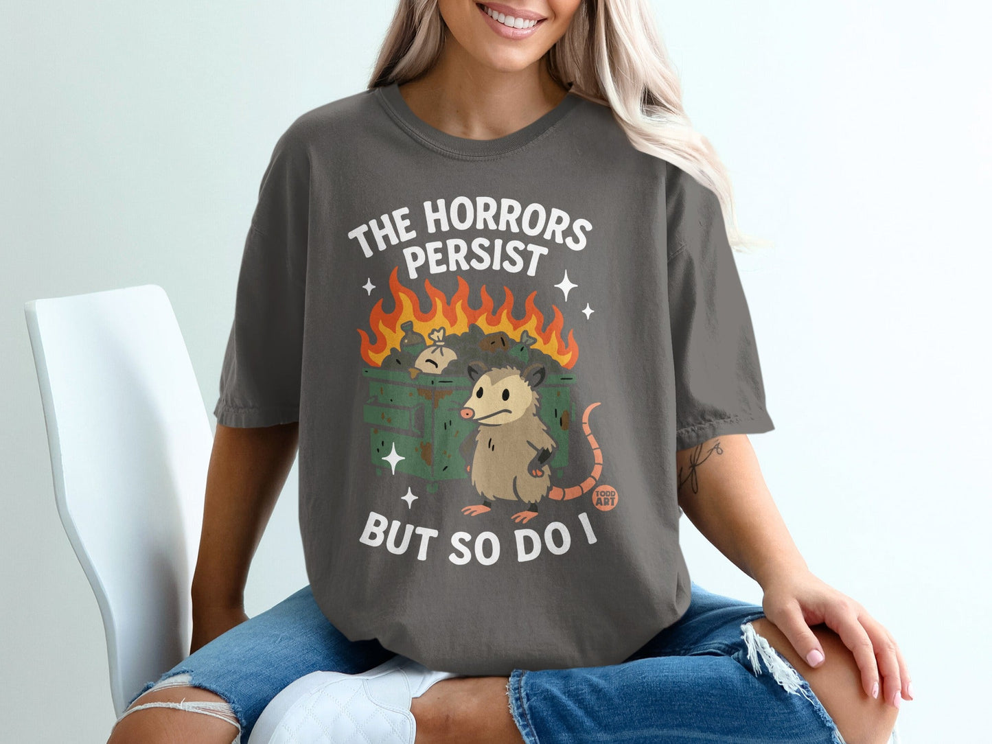 Dark gray t-shirt with opossum in trash and text "The horrors persist but so do I".